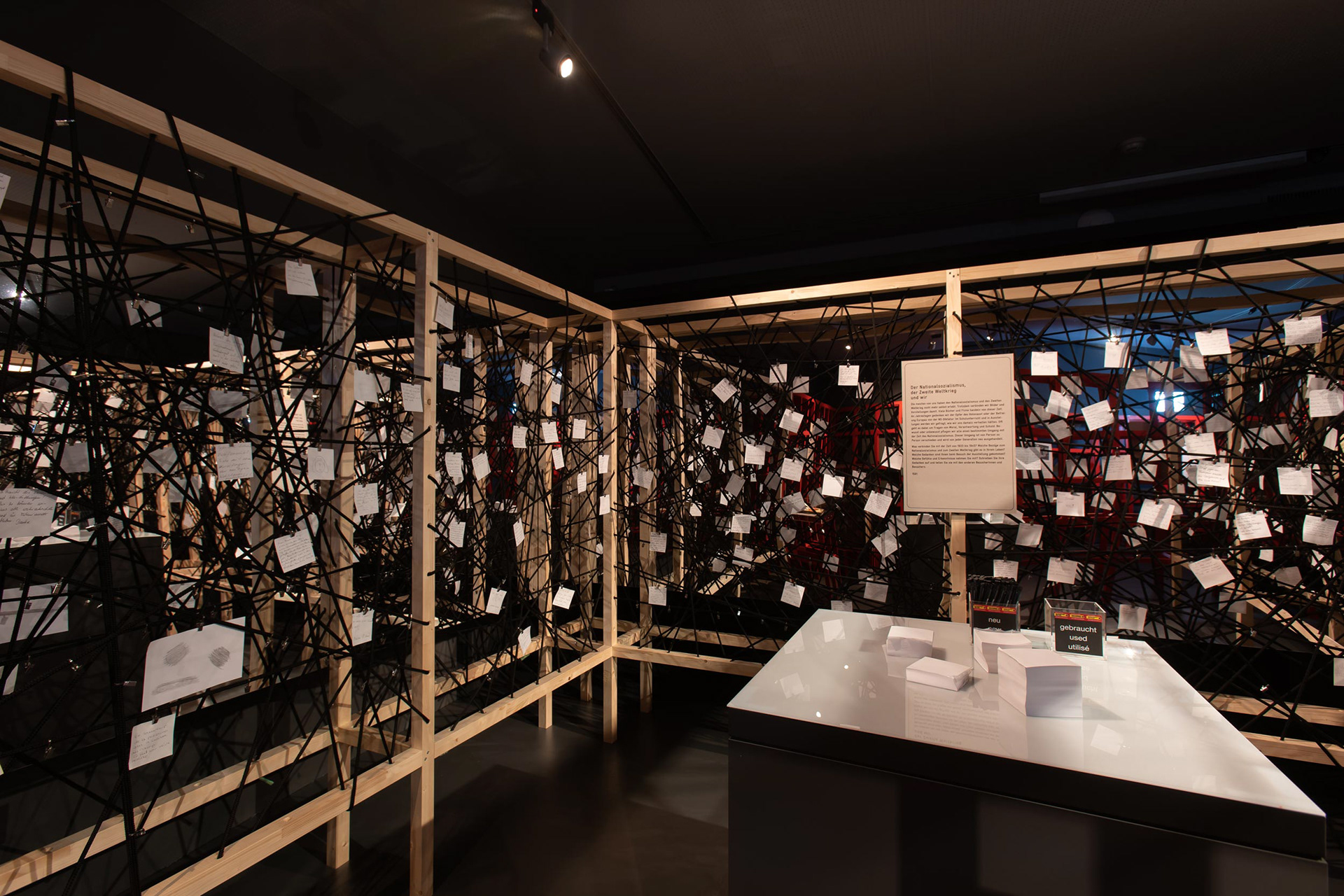 Writing station in the middle of the wooden racks. In the background of the spanned ropes there is a mirror film, which has the effect of not only reflecting the paper notes, but also the visitor himself.
