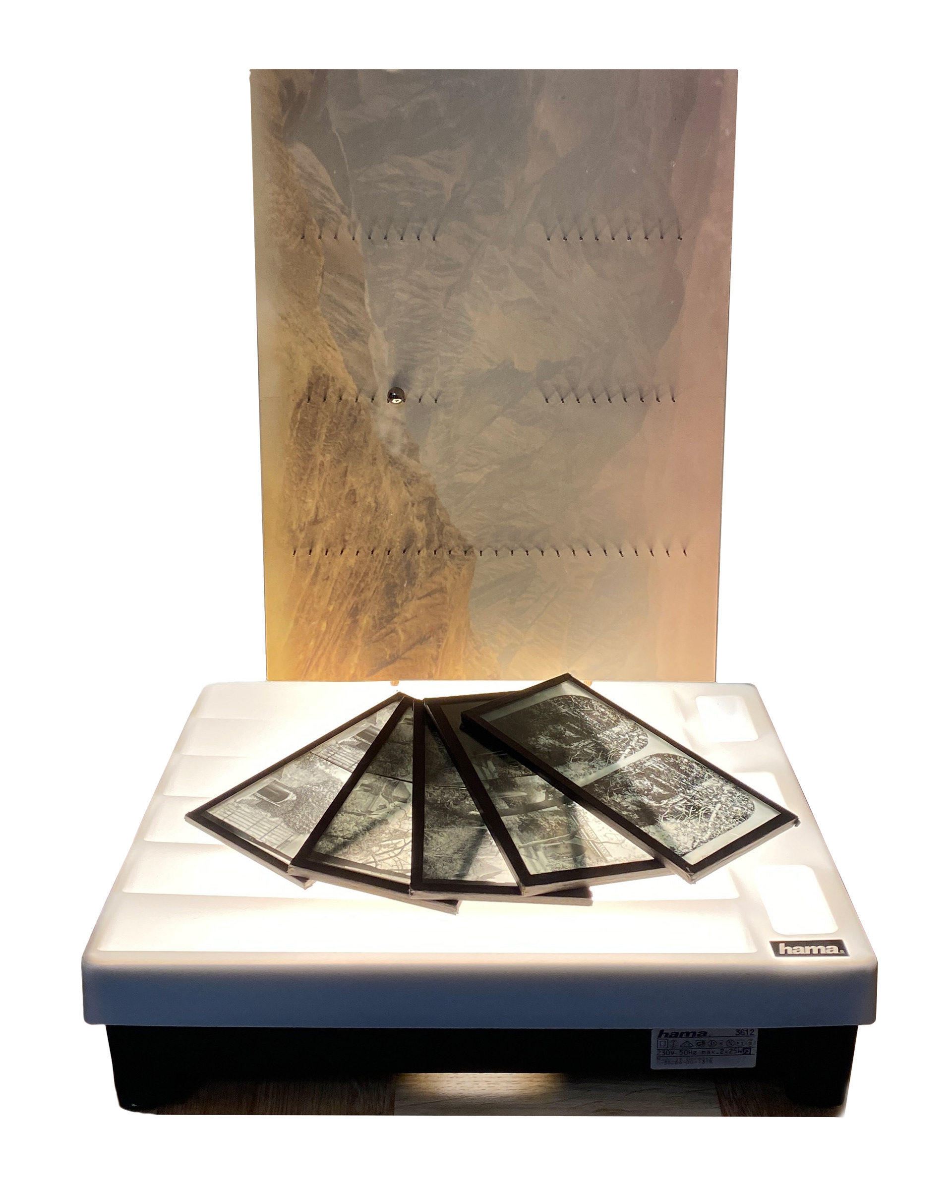 Art installation comprising of early 20th century glass photographic plates of landscapes, resting on an illuminated 70's slide viewer light-box along with a vertically mounted colour aerial photo of mountainous desert as background.