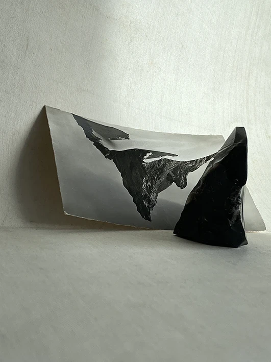 Still life photo of a black and white postcard depicting Zermatt mountain peak upside down and a triangular shaped piece of obsidian against a canvas textured background