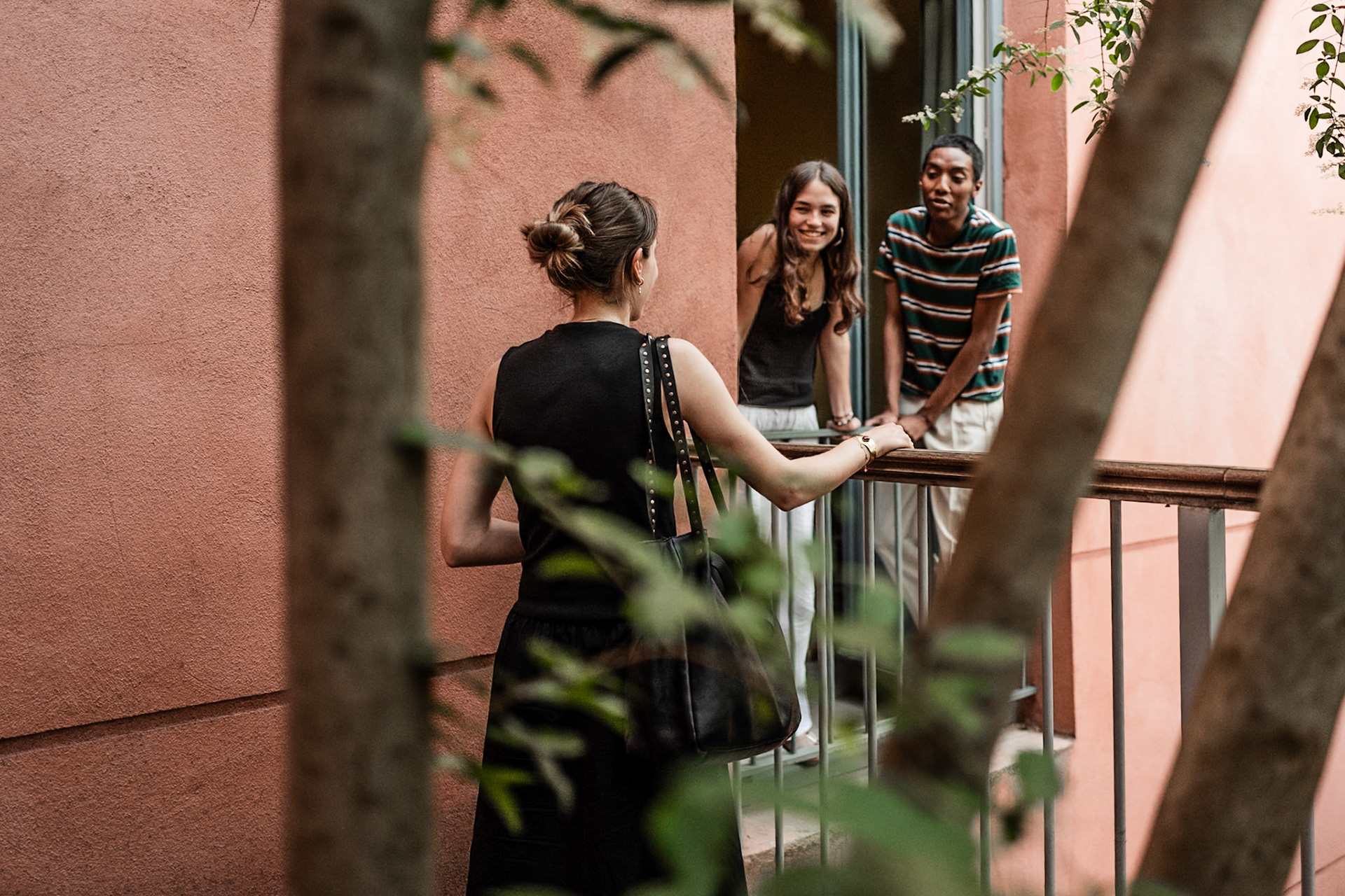 A candid scene of three people sharing a warm interaction between the terrasse and a balcony of a room, framed by trees and a soft light. A genuine moment of connection and everyday life. Picture taken the 2026-05-27©Aurora Ghigino