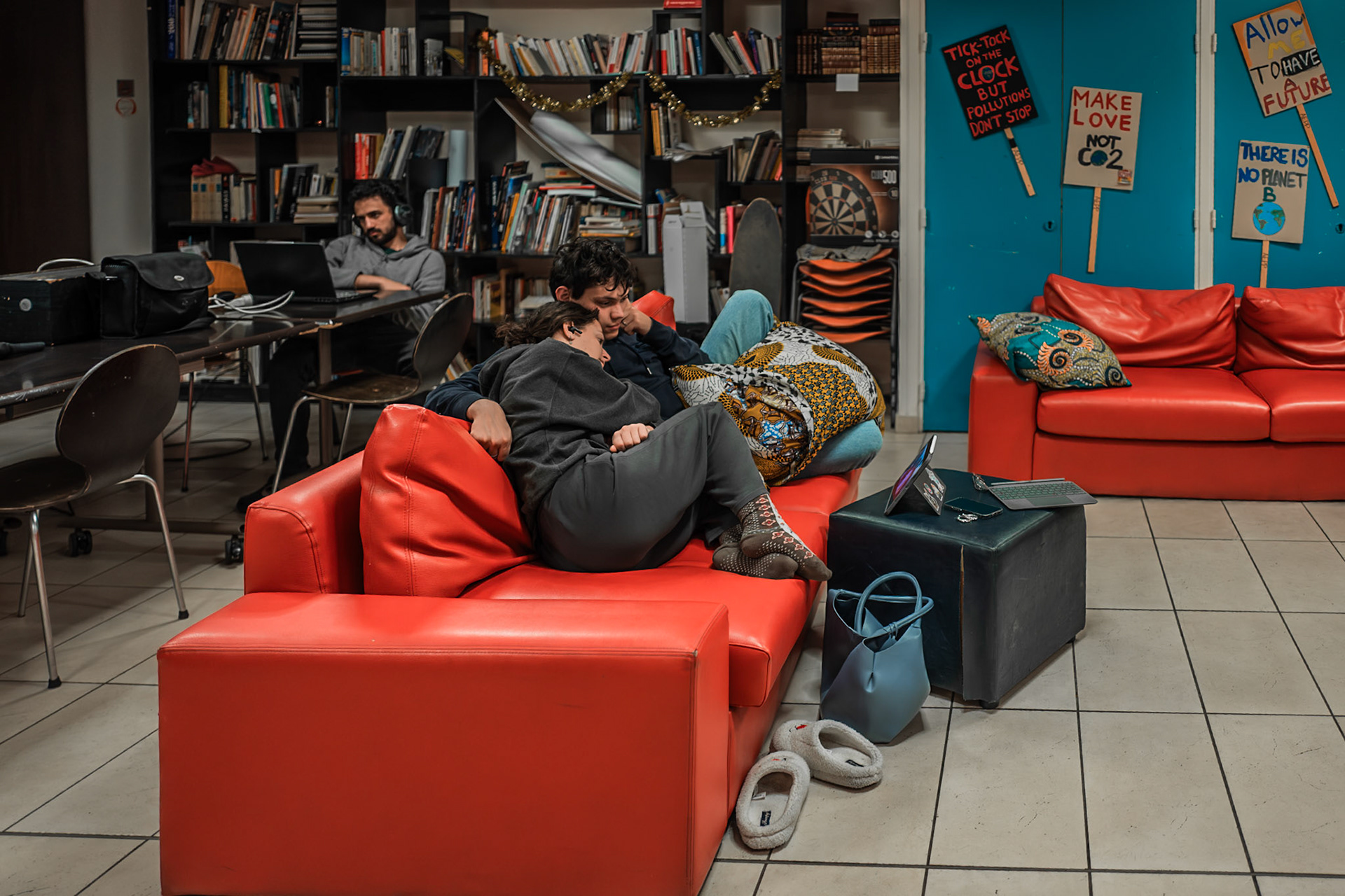 A young couple, Gaspard and Célimène, sit together in the main hall watching a film on a tablet, absorbed in their own private world and seemingly detached from their surroundings. In the background, a solitary student watches the same film alone. The contrast between the two scenes highlights different experiences of everyday life within the residence, revealing how shared spaces can hold both intimacy and solitude at a single glance. Picture taken the 2026-04-22©Aurora Ghigino