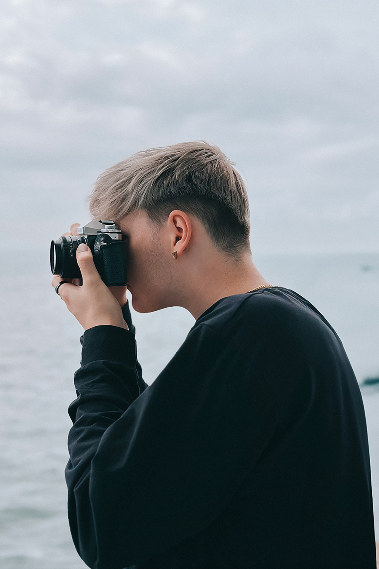 An image of Tom. He is looking through the viewfinder of a vintage 35mm SLR camera. You can see the ocean in the background.