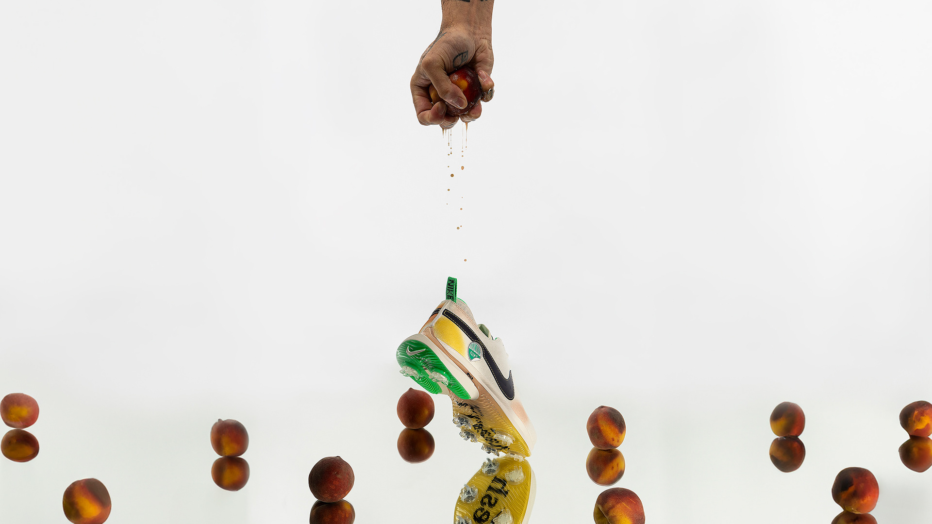 An image showing a Nike Air Zoom Victory Tour III NRG M23 Phantom/Gridiron/Midwest Gold (designed to resemble a peach) on a mirror with the heel in the air surrounded by peaches. A tattooed hand is squeezing a peach above the shoe and the juice is captured dripping.