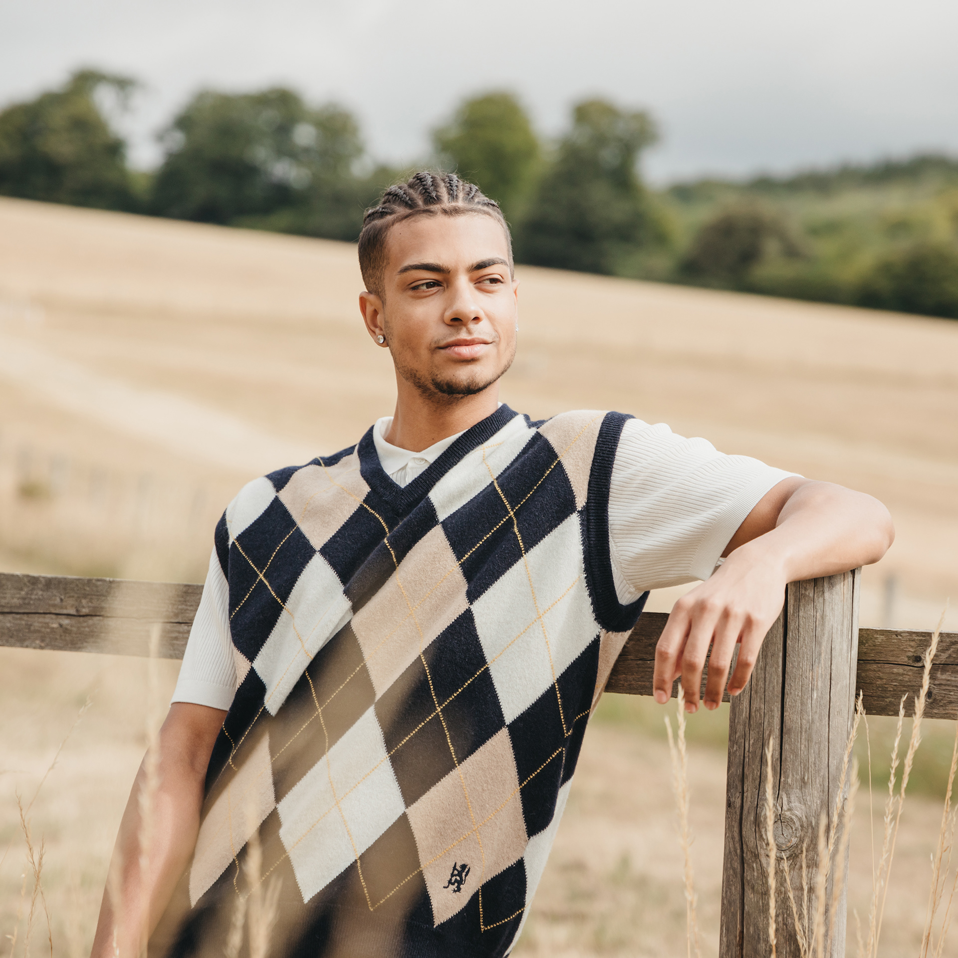 An example of Tom's Fashion and Editorial work. It shows a male subject on a warm summers day wearing a navy, beige and white argyle knitted vest by Pringle of Scotland. The subject is leant casually against a wooden fence and is looking to the right of the frame.