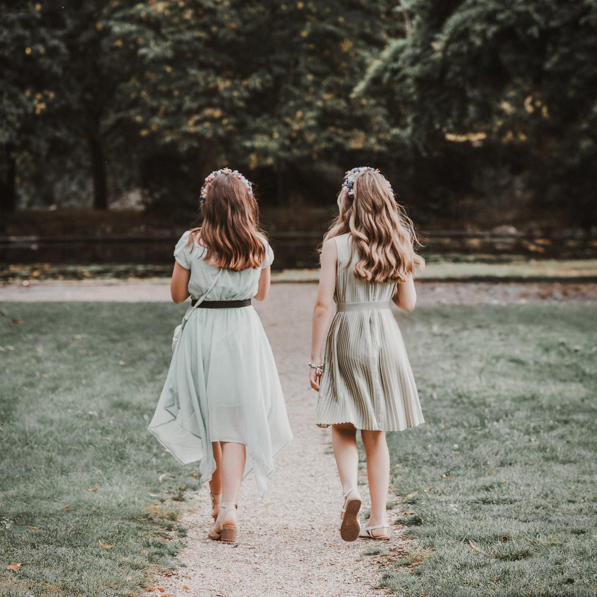 An example of Tom's Wedding and Event photography. The image shows two young girls walking away fro the camera. They are dressed in soft green dresses and are wearing flower crowns in their hair. The image has a nostalgic film aesthetic.