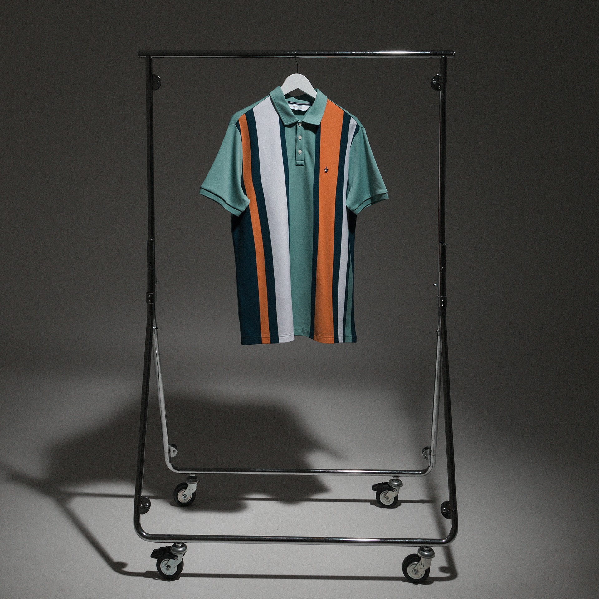 An example of Tom's Product and E-Commerce imagery. The image shows a polo shirt by Original Penguin in collaboration with TRENDYGOLF. The shirt is teal, navy, orange and white in a chunky vertical striped pattern. The item is hung with a white hanger on a small silver clothes rail and is lit by a single diffused spot light from above. This is creating soft shadowing across the white floor and backdrop.