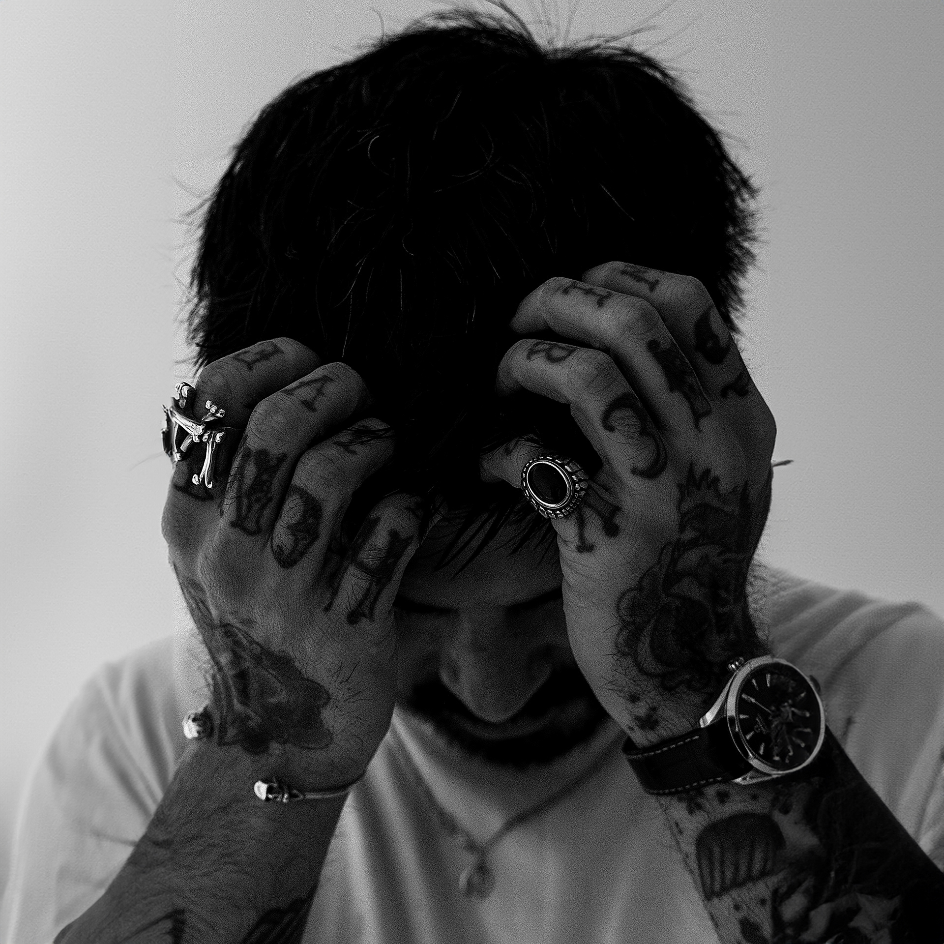 An example of a portrait by Tom. The image is a grainy black and white portrait of a dark haired man with tattoos on his hands and arms. He wears chunky jewellery including a silver skull band and watch with a leather strap. He wears a plain white T-shirt with a silver necklace visible. The subject is looking down and his head is in his hands. This image would serve well as an album cover.
