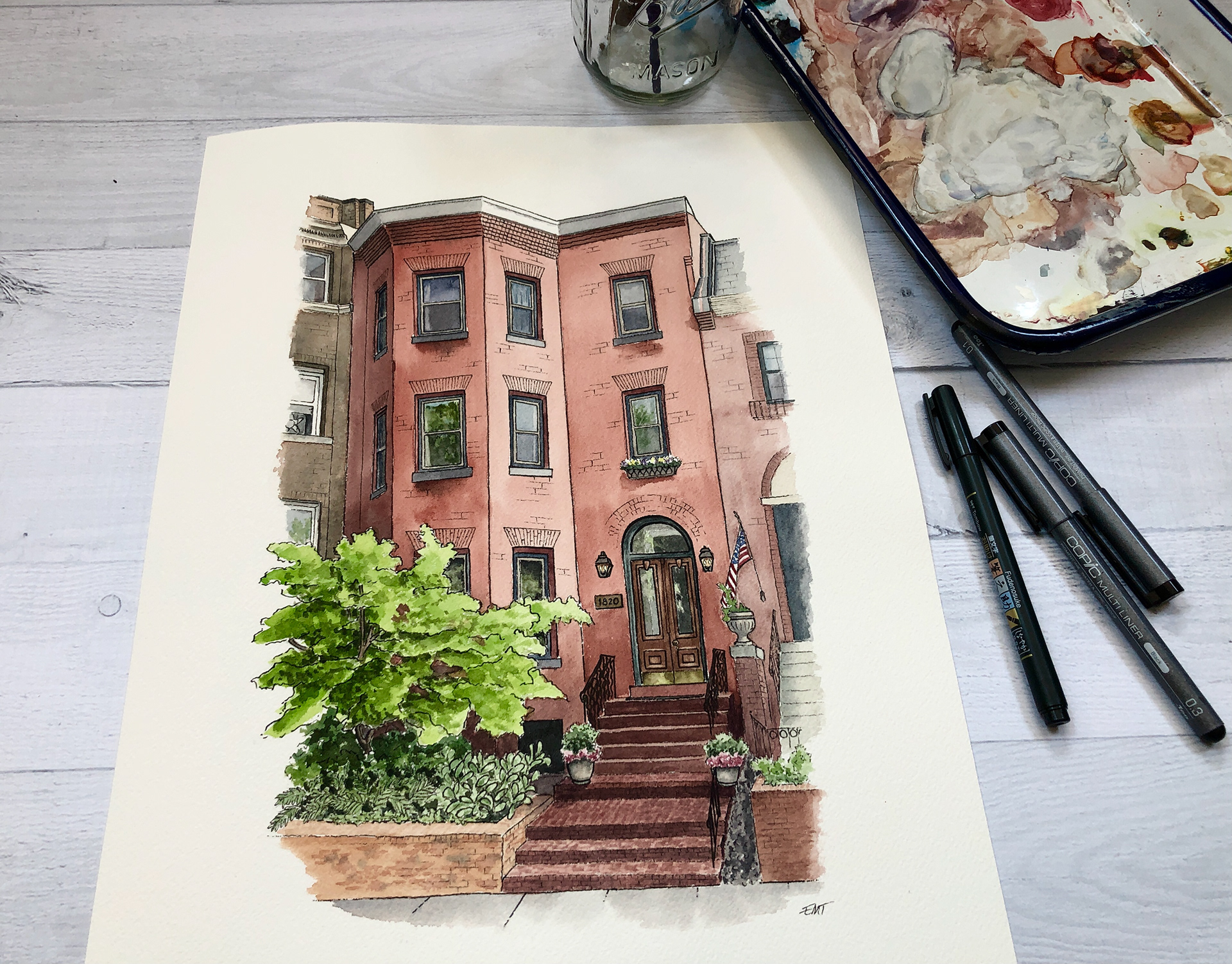 Pen and watercolor artwork of a red brick row house, alongside a paint palette, pens, and water jar