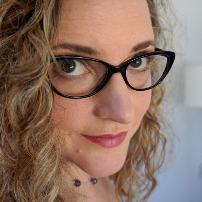 A close-up headshot of a woman with fair skin and curly, shoulder-length blond hair. She is wearing large, black-rimmed glasses, which highlight her green eyes. She has a neutral expression with a slight smile, and she's looking directly at the camera. The background is light-colored and out of focus, emphasizing her as the subject. She is wearing a dark top and a necklace with purple beads.
