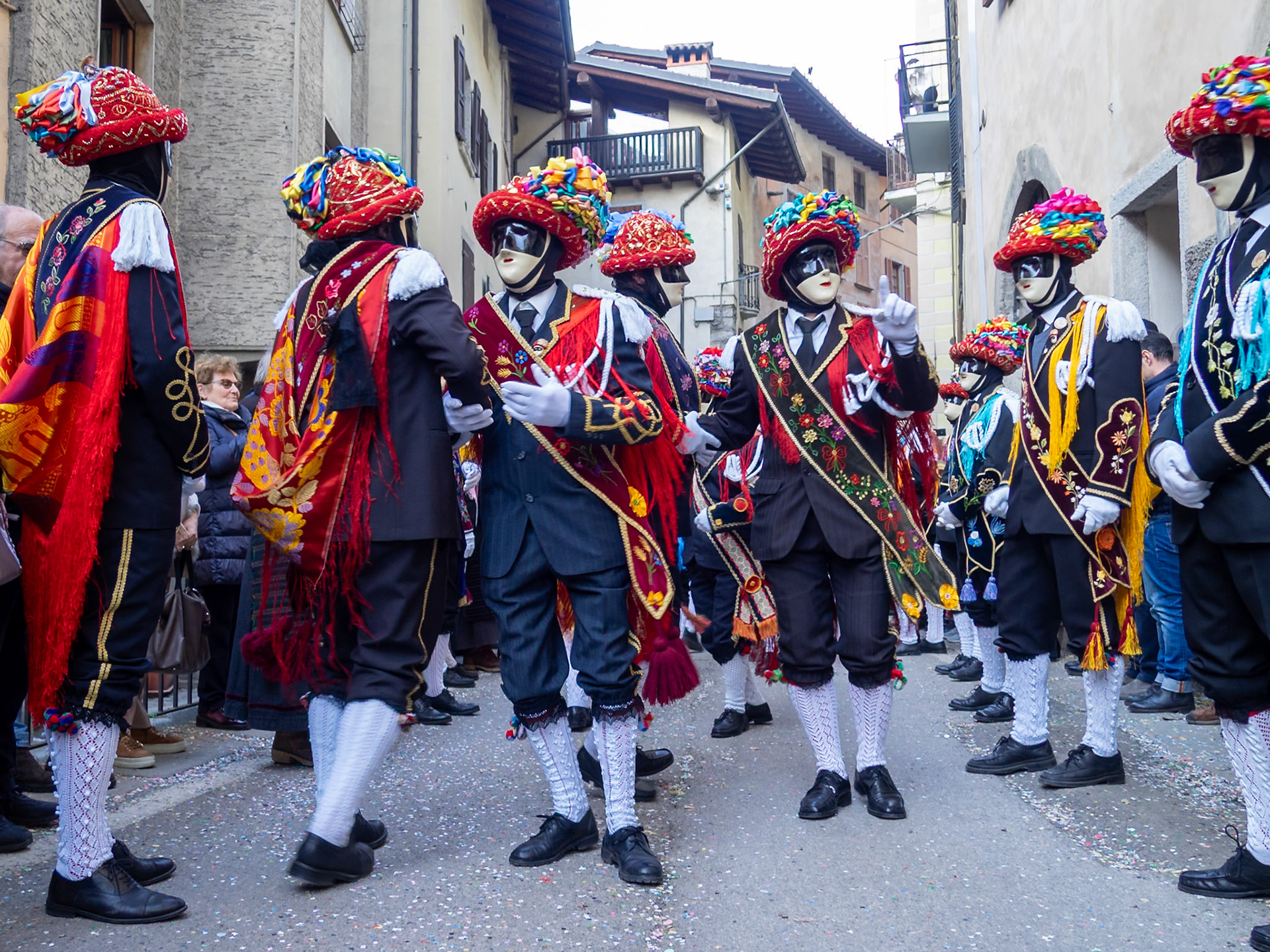 Balari dancing in Bagolino streets during Carnival, wearing the traditional costume with white knitted socks, black dress, colorful shawl over the back, face covered in a ivory and black mask, and head under a felt hat covered in red ribbon, with gold jewelry and multicolored ribbons forming a bow