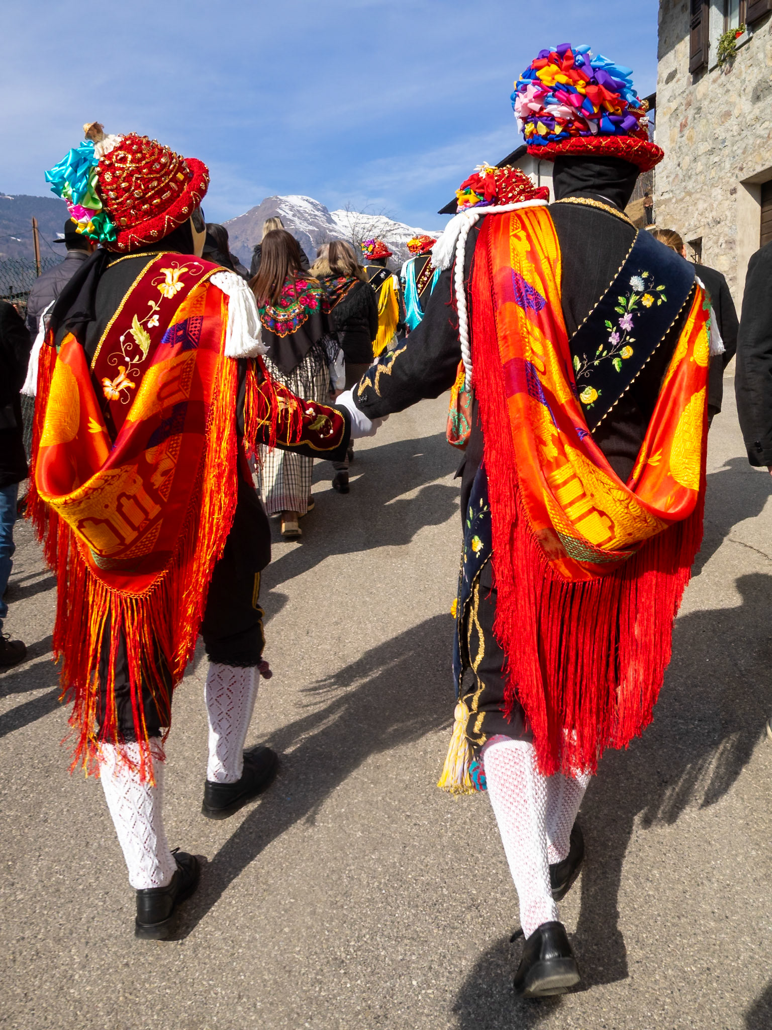 Balari walking in Bagolino streets during Carnival, wearing the traditional costume with face covered in a ivory and black mask, and head under a felt hat covered in red ribbon, with gold jewelry and multicolored ribbons forming a bow
