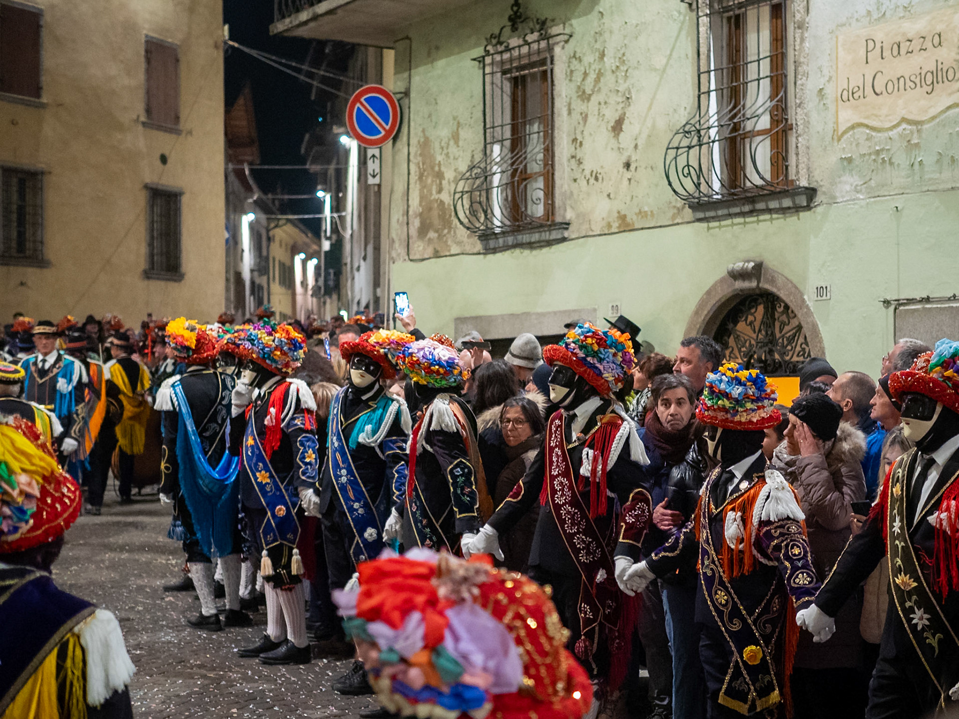 Balari dancing, at night, in Bagolino streets during Carnival celebrations closure, wearing the traditional costume with white knitted socks, black dress, colorful shawl over the back, face covered in a ivory and black mask, and head under a felt hat covered in red ribbon, with gold jewelry and multicolored ribbons forming a bow