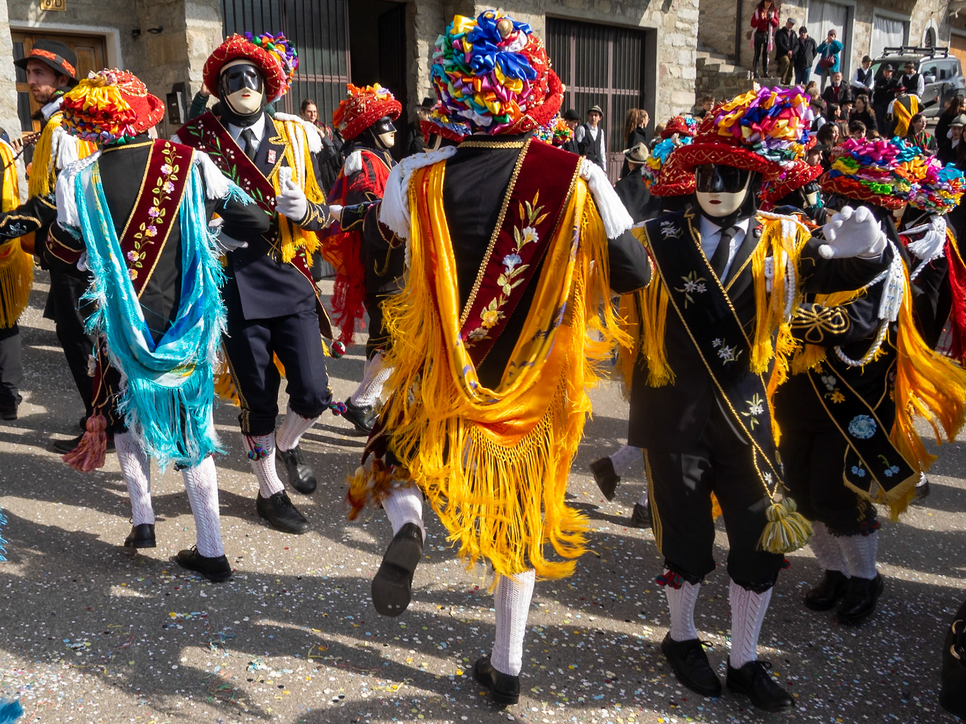 Balari dancing in Bagolino streets during Carnival, wearing the traditional costume with white knitted socks, black dress, colorful shawl over the back, face covered in a ivory and black mask, and head under a felt hat covered in red ribbon, with gold jewelry and multicolored ribbons forming a bow