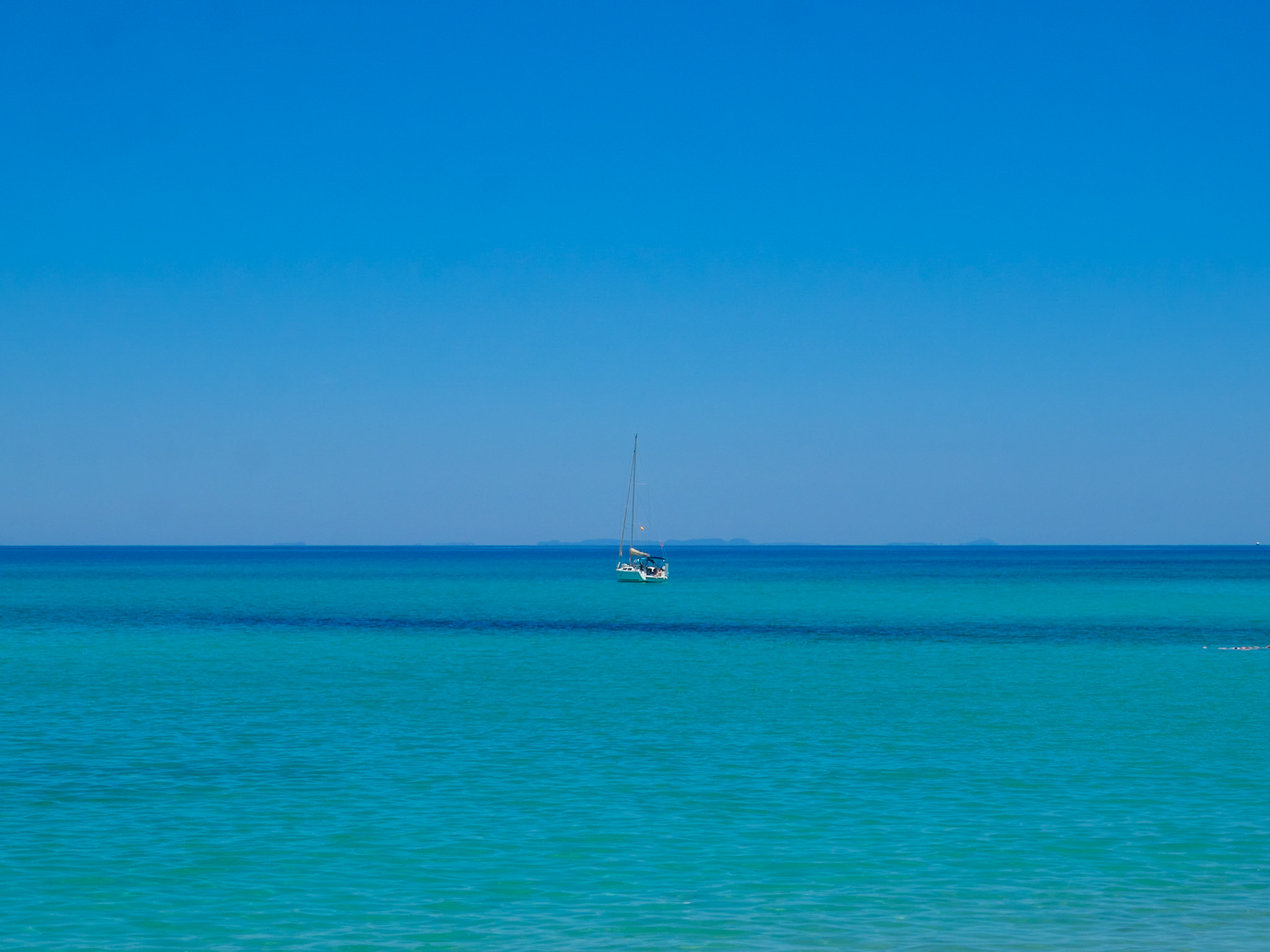 Sailboat in the horizon line between turquoise waters and clear blue sky