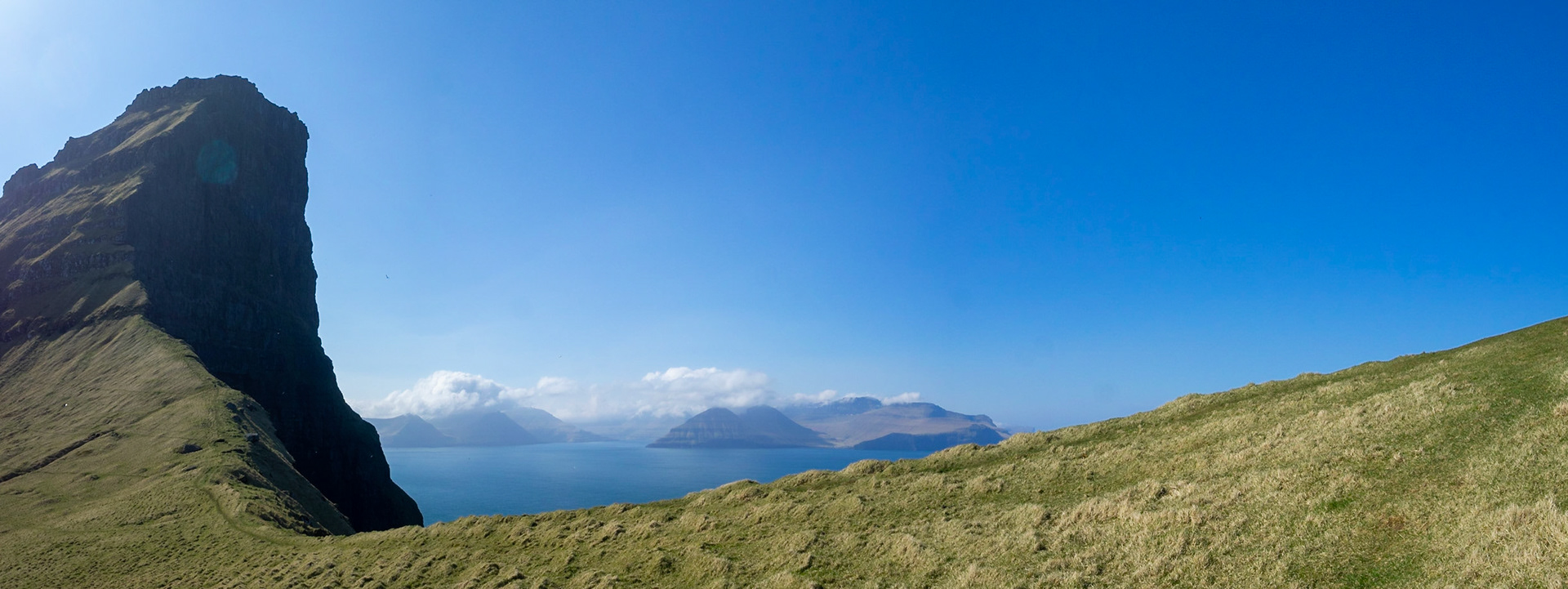 Panorama of Kallur lighthouse hiking path by the cliffs edge, in north Kalsoy, with Eysturoy island in the horizon