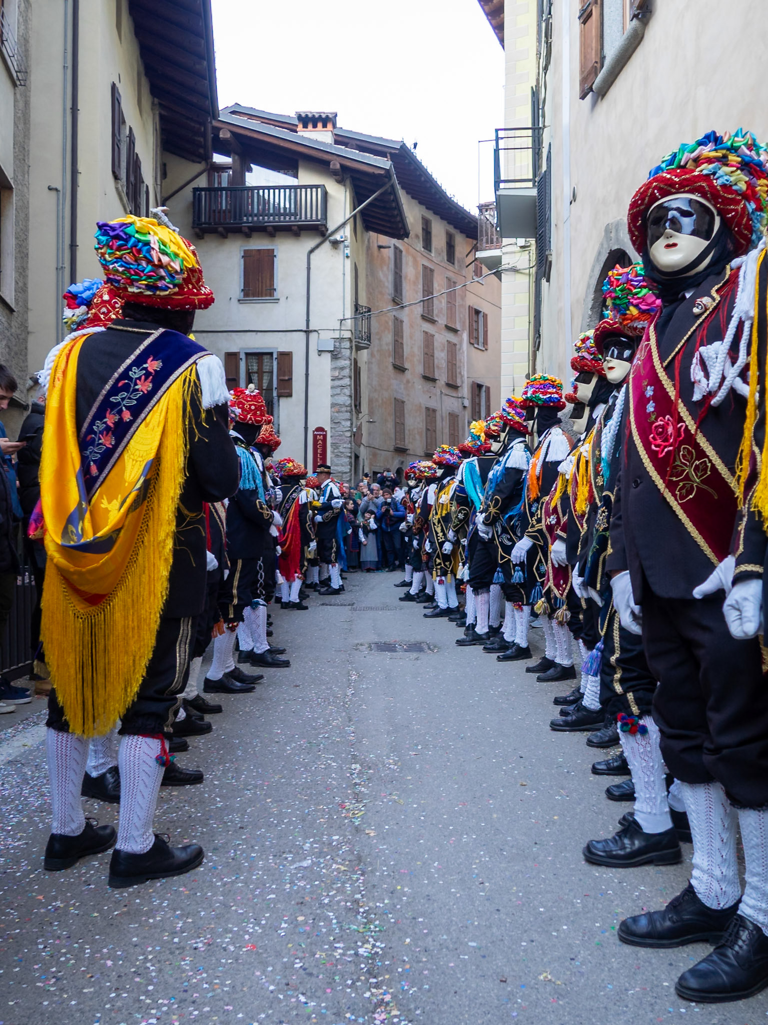 Balari dancing in Bagolino streets during Carnival, wearing the traditional costume with white knitted socks, black dress, colorful shawl over the back, face covered in a ivory and black mask, and head under a felt hat covered in red ribbon, with gold jewelry and multicolored ribbons forming a bow