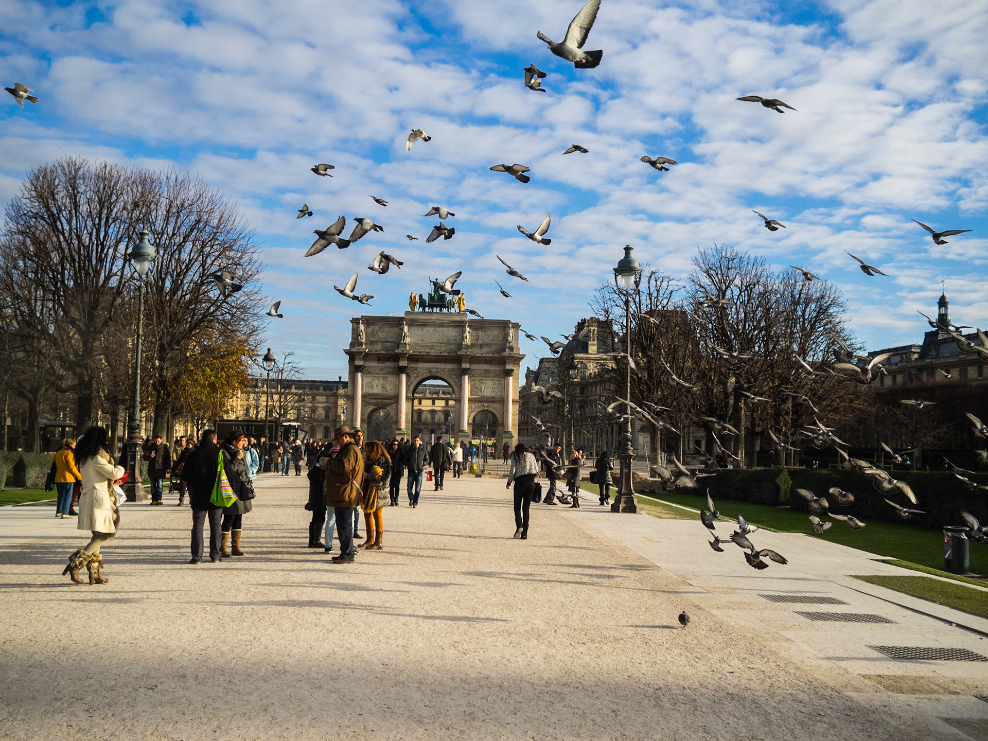 Pigeons fly by tourists close to Carrousel du Louvre arch