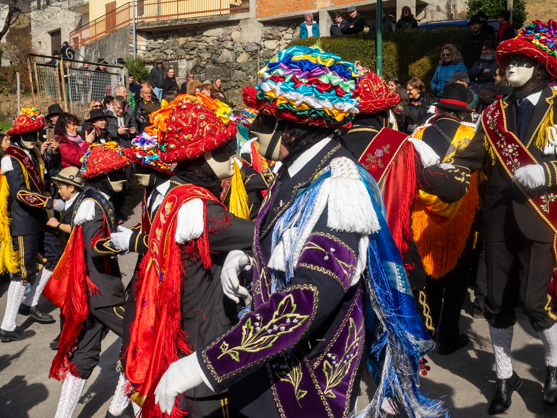 Balari dancing in Bagolino streets during Carnival, wearing the traditional costume with white knitted socks, black dress, colorful shawl over the back, face covered in a ivory and black mask, and head under a felt hat covered in red ribbon, with gold jewelry and multicolored ribbons forming a bow