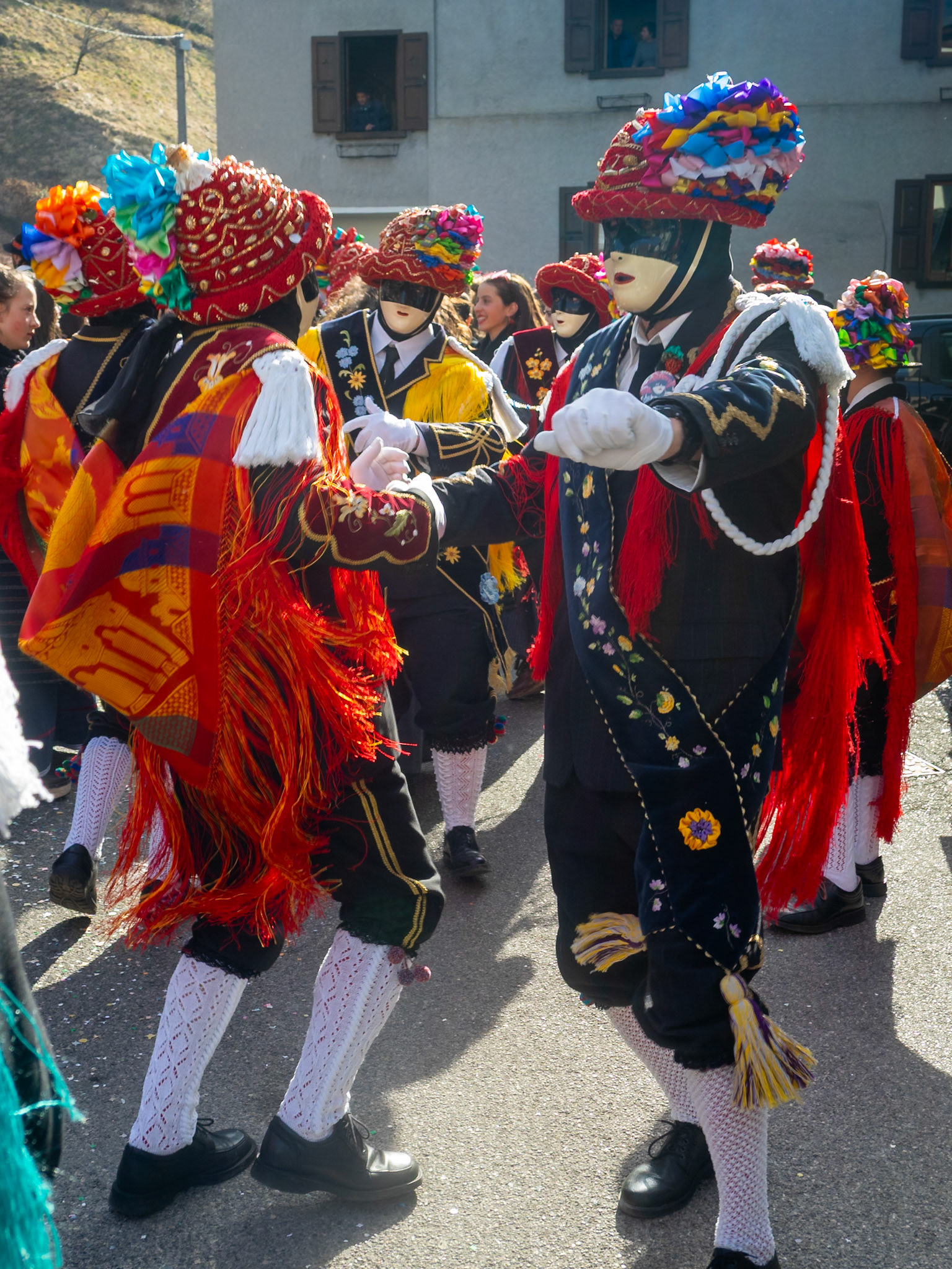 Balari dancing in Bagolino streets during Carnival, wearing the traditional costume with white knitted socks, black dress, colorful shawl over the back, face covered in a ivory and black mask, and head under a felt hat covered in red ribbon, with gold jewelry and multicolored ribbons forming a bow