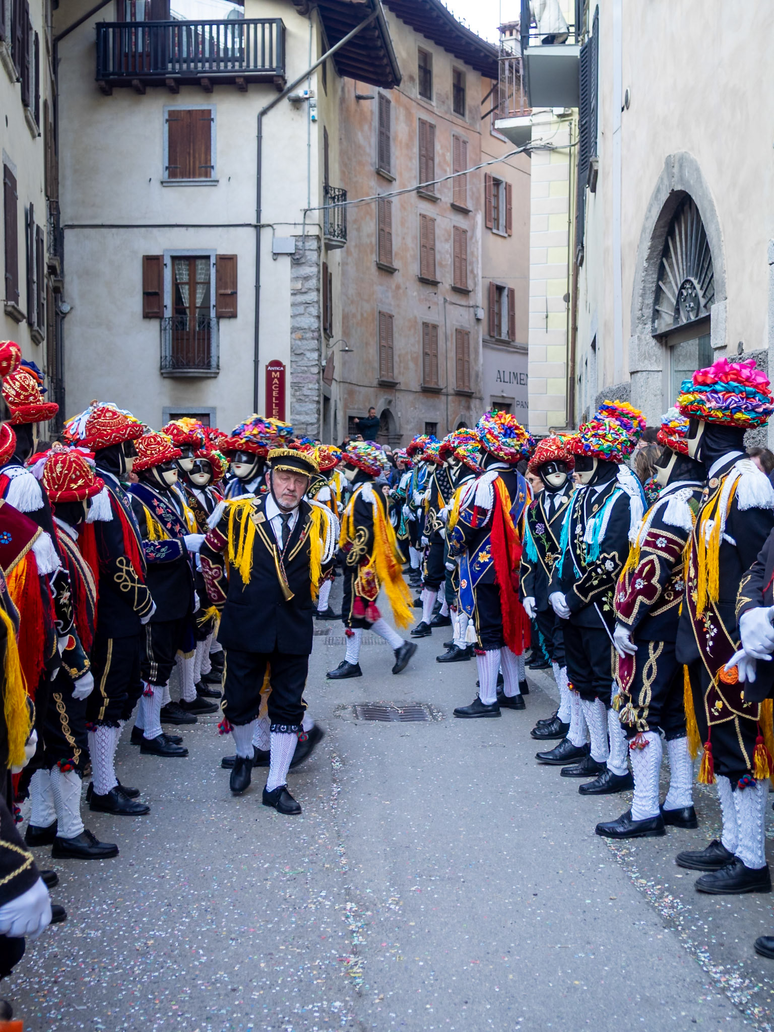 Balari dancing in Bagolino streets during Carnival, wearing the traditional costume with white knitted socks, black dress, colorful shawl over the back, face covered in a ivory and black mask, and head under a felt hat covered in red ribbon, with gold jewelry and multicolored ribbons forming a bow