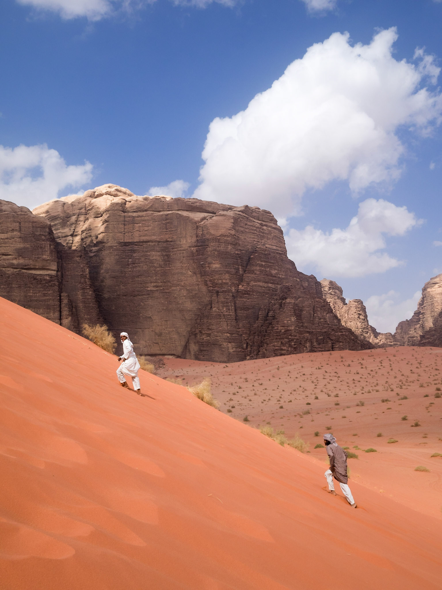 Two men dressed in Arabian costume climb up the red sand dunes of the Wadi Rum desert