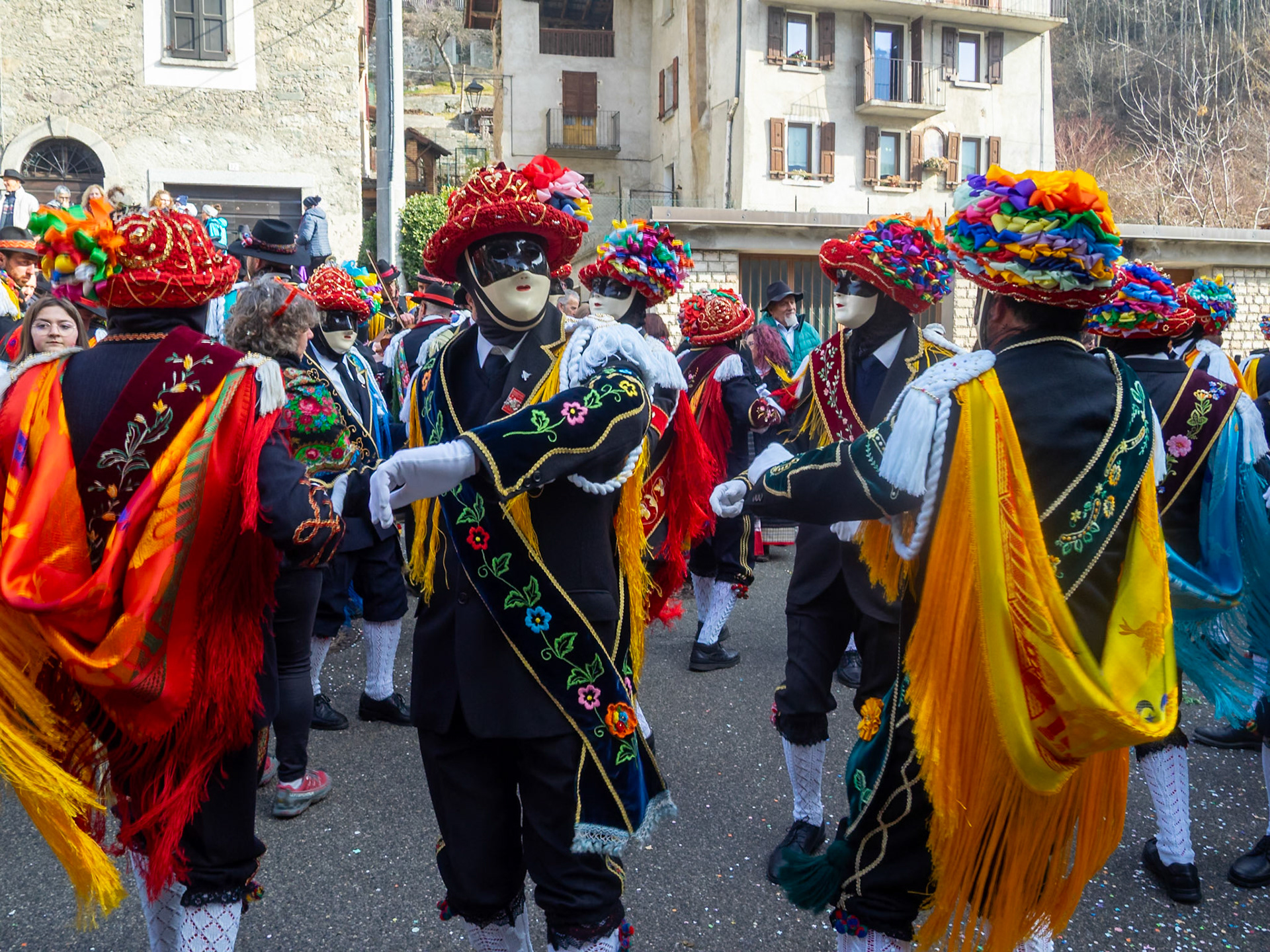 Balari dancing in Bagolino streets during Carnival, wearing the traditional costume with white knitted socks, black dress, colorful shawl over the back, face covered in a ivory and black mask, and head under a felt hat covered in red ribbon, with gold jewelry and multicolored ribbons forming a bow