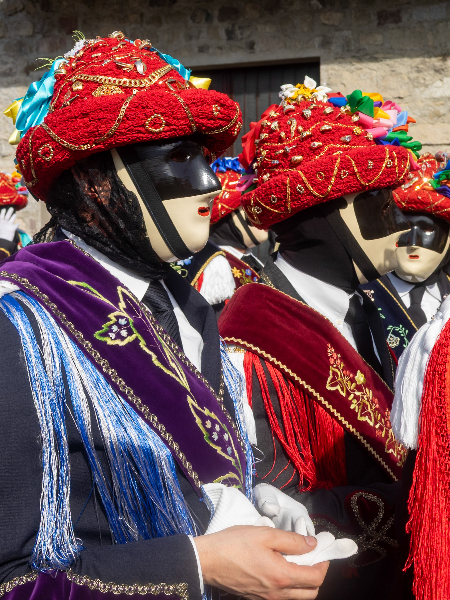 Bagolino Carnival Balari portrait, face covered by the expressionless ivory and black colored mask, head under a felt hat, covered in red ribbon, gold jewelry and colorful ribbons, wearing a black dress  with a silk band with flowers embroidery and white gloves