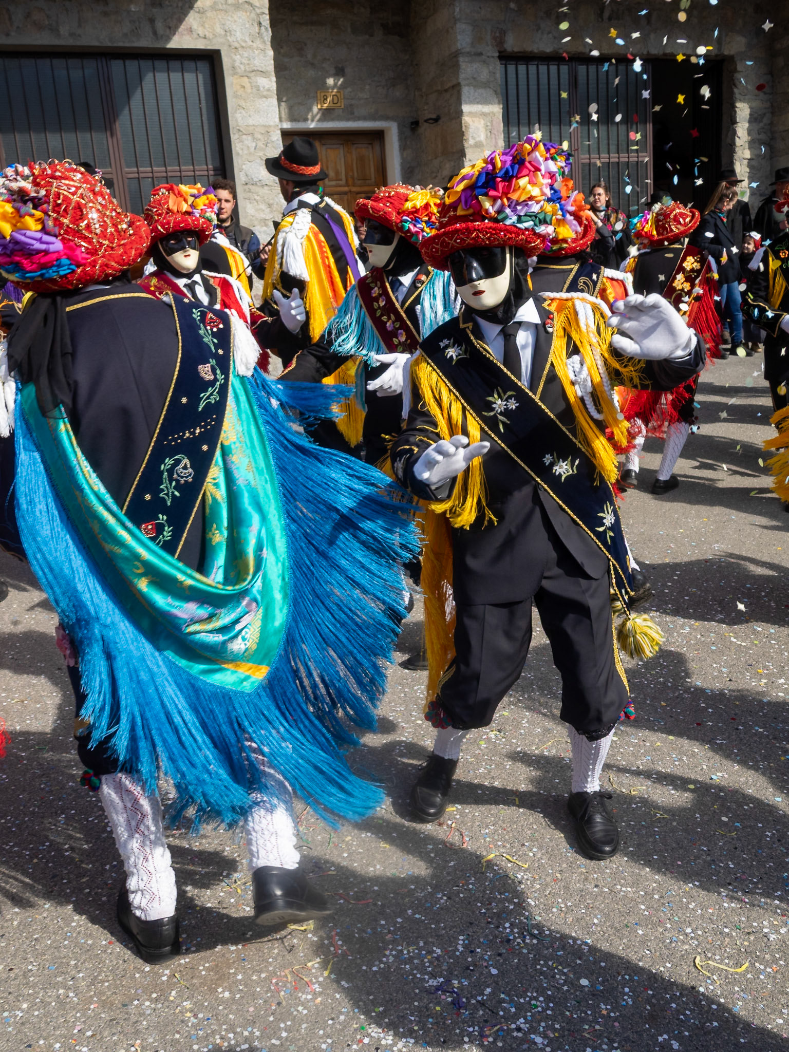 Balari dancing in Bagolino streets during Carnival, wearing the traditional costume with white knitted socks, black dress, colorful shawl over the back, face covered in a ivory and black mask, and head under a felt hat covered in red ribbon, with gold jewelry and multicolored ribbons forming a bow