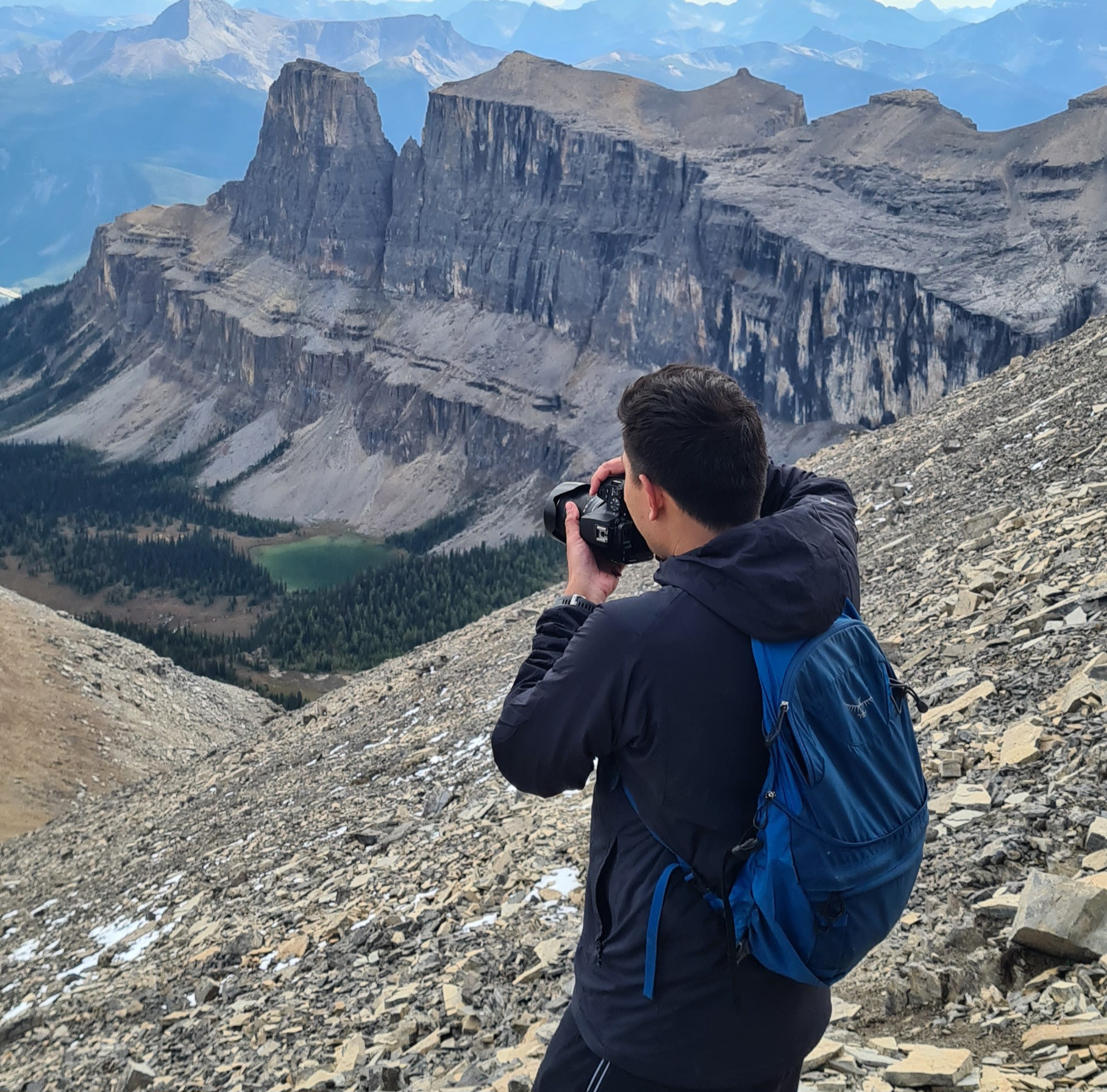 Matt stands turned away on the side of a mountain, lining his camera up to take a photo of other mountain ranges.