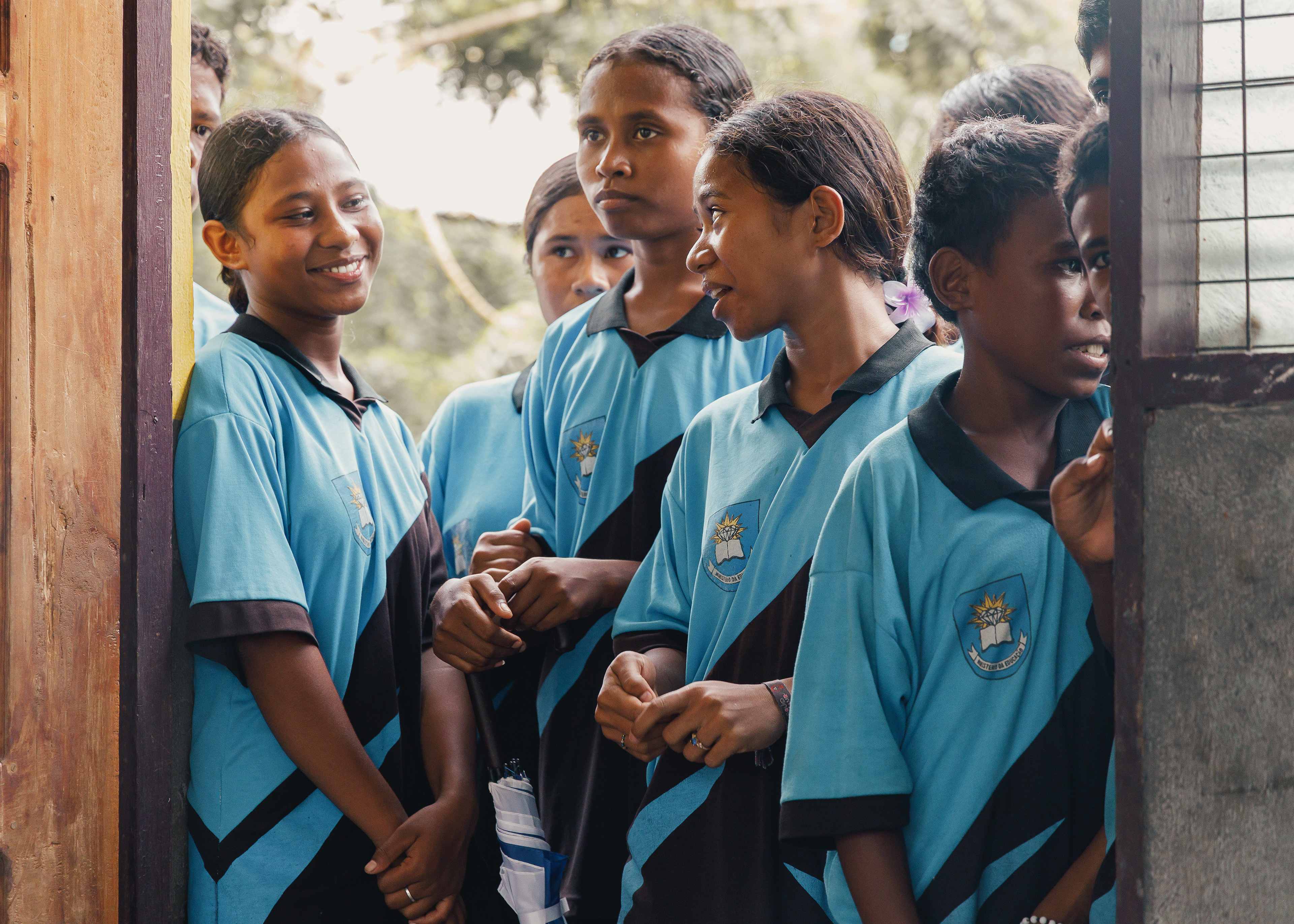 A few students are standing at the door, ready to enter the classroom, their expressions reflecting a strong spirit and eagerness to learn, despite the limitations of books, learning equipment, libraries, computers, and labs.