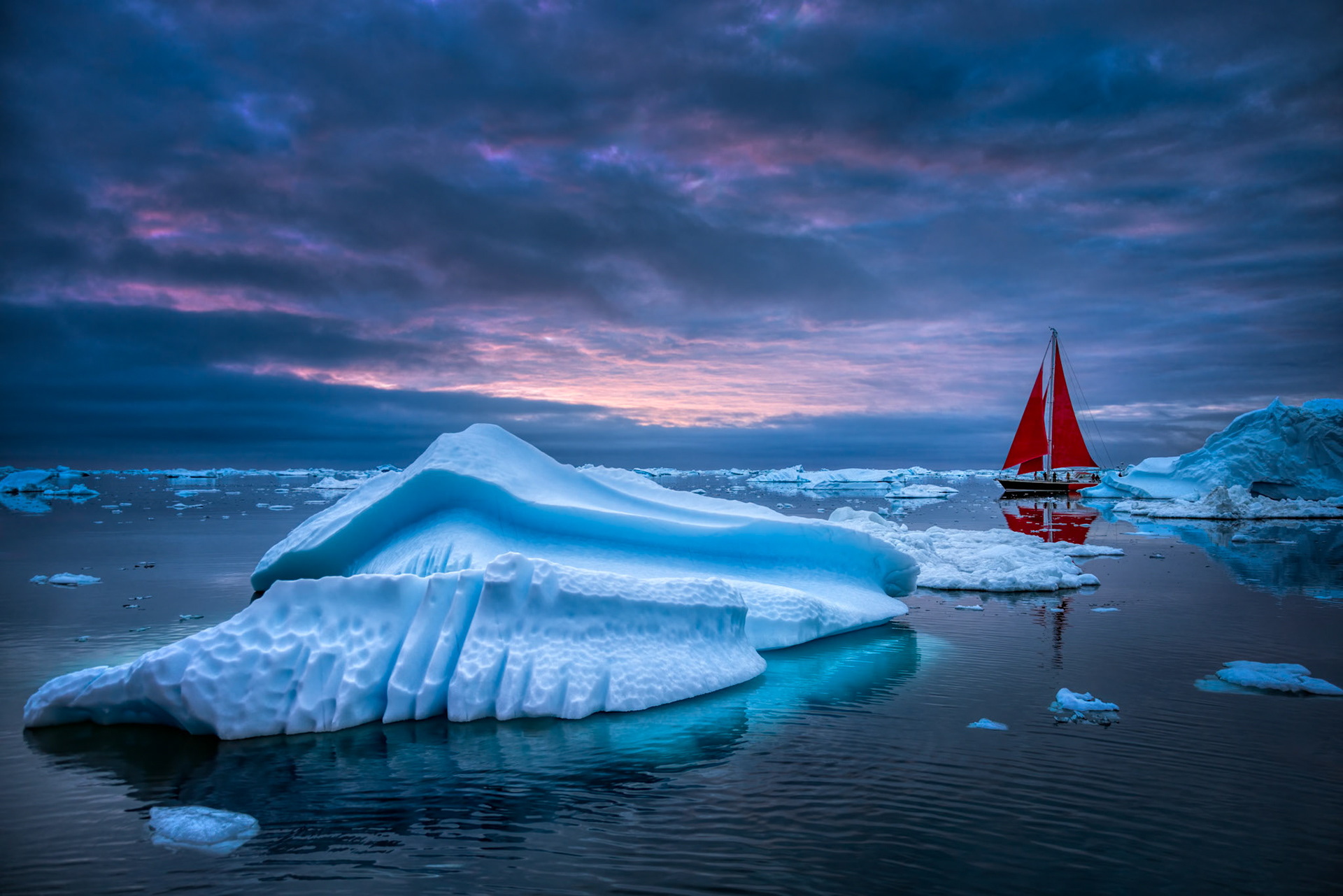 RED AGAINST THE COLD  |  A lone sailboat with brilliant red sails glides through a maze of icebergs along Greenland's rugged coast, providing a striking contrast against the cool blues and silvers of the Arctic. The sculpted iceberg in the foreground appears almost carved by hand, its smooth curves and intricate textures shaped over centuries by wind, water, and the relentless forces of nature.Photographed during the fleeting light of an Arctic summer evening, the scene captures both the serenity and scale of one of the planet's last great wildernesses. The small vessel serves as a reminder of humanity's place within this immense landscape, while the glowing sails draw the eye toward the horizon where sea, ice, and sky merge into a palette of soft purples and blues. It is a moment of quiet exploration, where adventure and solitude coexist in one of the most remote corners of the world.