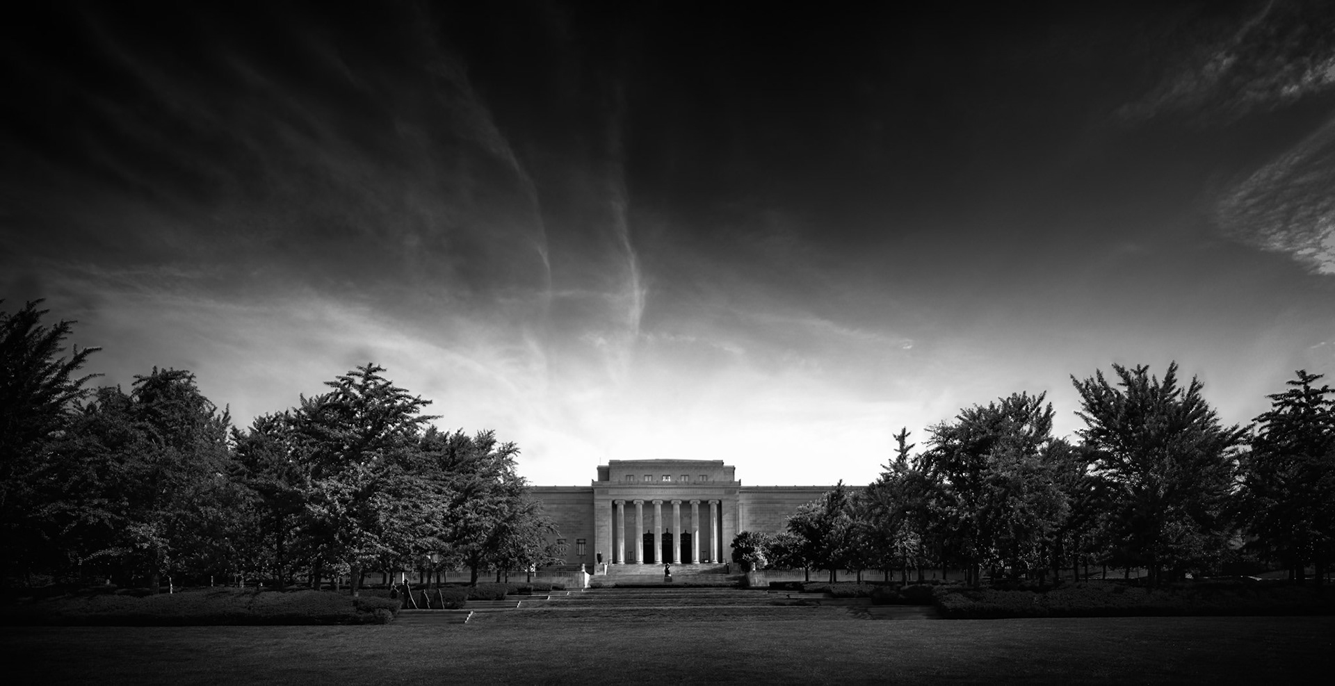 THE QUITE OF NELSON  |  Great museums are more than buildings. They are places where imagination, history, and human creativity come together. Standing before the Nelson Art Gallery in Kansas City, I was struck by the sense of permanence and quiet dignity that defines the institution.Rather than focus solely on the architecture, I chose a wider perspective that emphasized the relationship between the museum and its surroundings. The symmetrical rows of trees draw the eye toward the classical façade, while the dramatic sky above adds a sense of scale and atmosphere. The building feels almost ceremonial, standing as a beacon of culture amid an ever changing world.The monochrome treatment removes distractions and emphasizes form, light, and texture. The museum's clean lines and imposing columns become timeless, while the streaking clouds overhead introduce movement and energy. Together they create a balance between stability and change, permanence and motion.Museums preserve the achievements of the past, but they also inspire future generations. In that sense, the Nelson is more than a repository of art. It is a monument to curiosity, creativity, and the enduring value of human expression.