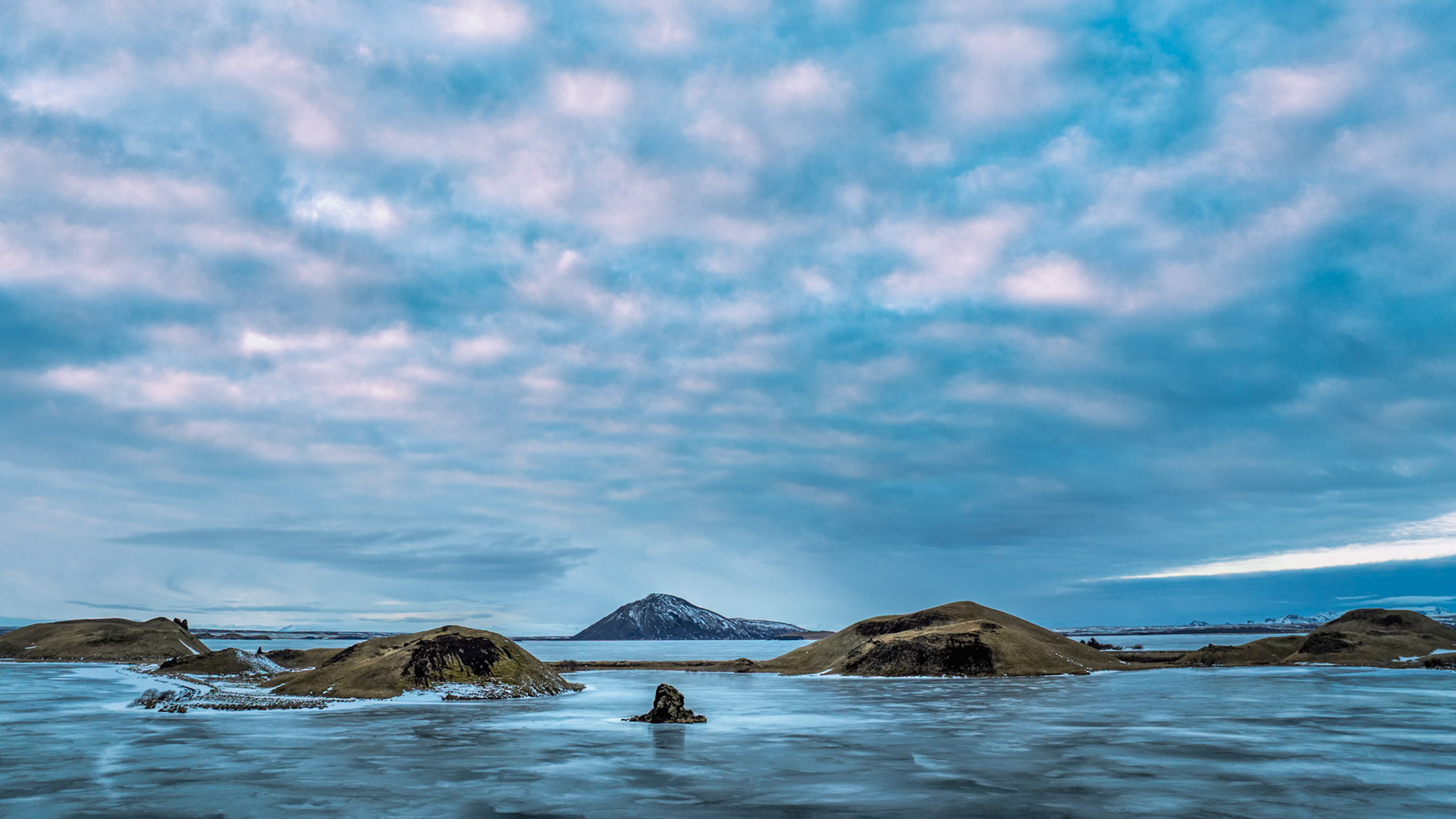 WHERE THE LAND WAITS  |  Winter settles gently across Lake Mývatn, transforming one of Iceland's most dynamic landscapes into a study of stillness. The frozen surface stretches toward the horizon like brushed glass, interrupted only by volcanic islets that rise from the ice like ancient sentinels.