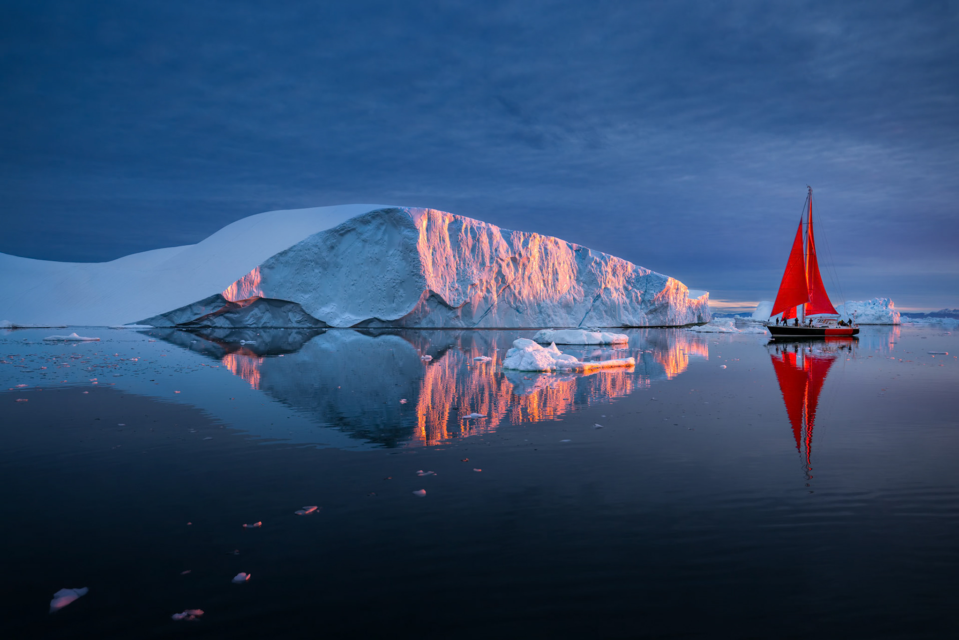 CRIMSON PASSAGE  |  This image captures one of those rare moments that photographers dream about but seldom experience. At 1:07 a.m. along the coast of Greenland, the midnight sun was hidden below the horizon, yet its light still found a way to illuminate the face of this massive iceberg in shades of pink and gold. The glow lasted only a few minutes before fading back into the cool blues of the Arctic night.What makes the scene even more extraordinary is the contrast between the immense iceberg and the small sailboat with its brilliant red sails. Together they create a sense of scale that is difficult to comprehend in person. The iceberg, sculpted over centuries within Greenland's ice sheet, reflects perfectly in the calm water, while the sailboat drifts quietly alongside one of nature's oldest creations.Of all the images I have made, this one remains my favorite. It combines everything that draws me to the Arctic: solitude, adventure, extraordinary light, and landscapes that seem almost impossible.