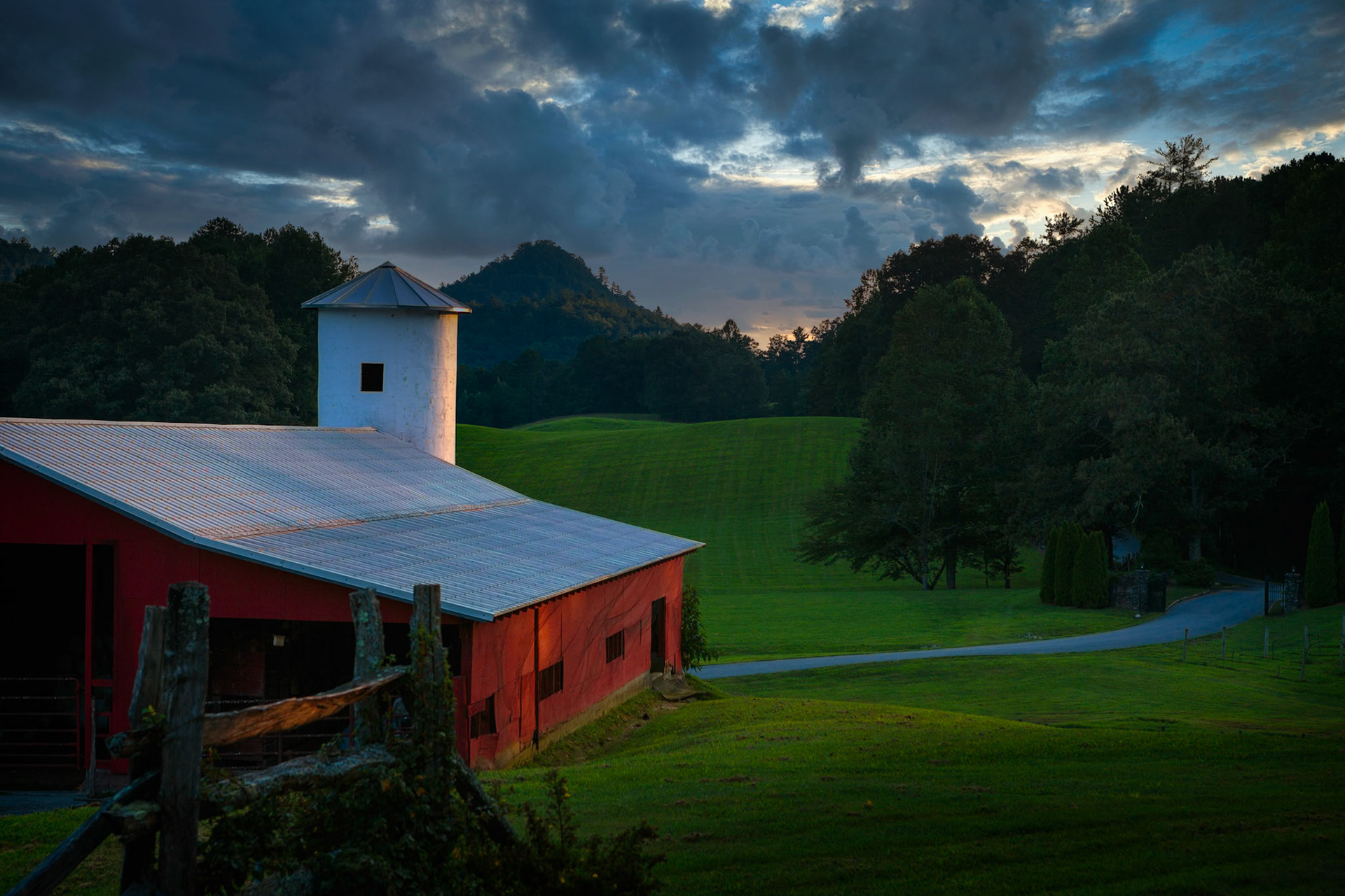 DUSK IN THE VALLEY  |  As evening settles across the hills near Lake Burton, the weathered red barn stands quietly against a landscape washed in deep greens and fading blue light. The last glow of the day lingers on the horizon, illuminating the rolling pasture and winding country road with a sense of calm that feels untouched by time.There is something comforting about rural scenes like this. The barn, sturdy and familiar, becomes more than a building. It is a symbol of tradition, resilience, and a slower pace of life. Surrounded by forested hills and gathering clouds, it anchors the landscape with quiet confidence.The photograph captures that fleeting moment between day and night when the world seems to exhale. The colors soften, the shadows deepen, and the countryside settles into silence. It is a reminder that some of life's most meaningful moments are not dramatic at all, but found in the peaceful beauty of simply being present.