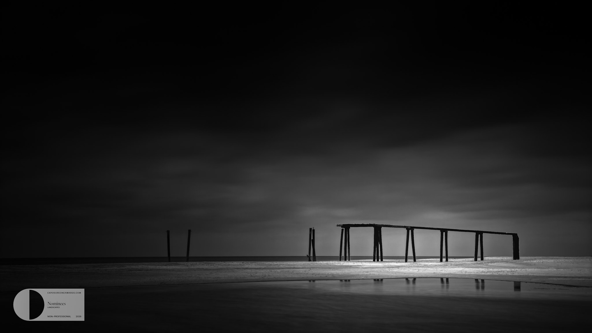 AFTER THE LAST TIDE  |  2026 EXPOSUREONE NOMINEE AWARD FOR LANDSCAPES  |  Not every photograph needs a dramatic subject. Sometimes a feeling is enough.On this quiet shoreline, what drew me in was not the structure itself, but the overwhelming sense of isolation it conveyed. The remains of an old pier stand alone against the sea and sky, disconnected from whatever purpose they once served. Time, storms, and tides have taken their toll, leaving only a fragment behind.The long exposure transformed the scene into something almost dreamlike. The water softened, the clouds stretched across the sky, and the world seemed to slow to a whisper. In that stillness, the weathered posts became less an object and more a symbol.We all experience moments of solitude. Sometimes they are peaceful, offering reflection and renewal. Other times they carry a sense of longing, reminding us of what once was. This abandoned structure seemed to embody both emotions at once.What remains standing is often more powerful than what has been lost.