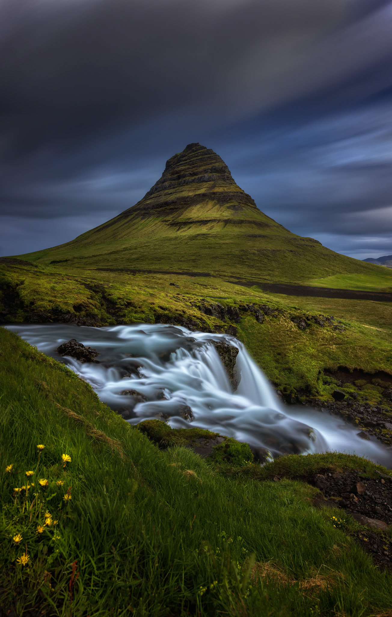 STORMLIGHT OVER KIRKJUFELL  |  Rising from the Icelandic landscape like a solitary sentinel, Kirkjufell stands beneath racing storm clouds while the waterfall below traces a path of silk through the foreground. For a brief moment, shifting light broke through the darkness, revealing the mountain's unmistakable form.