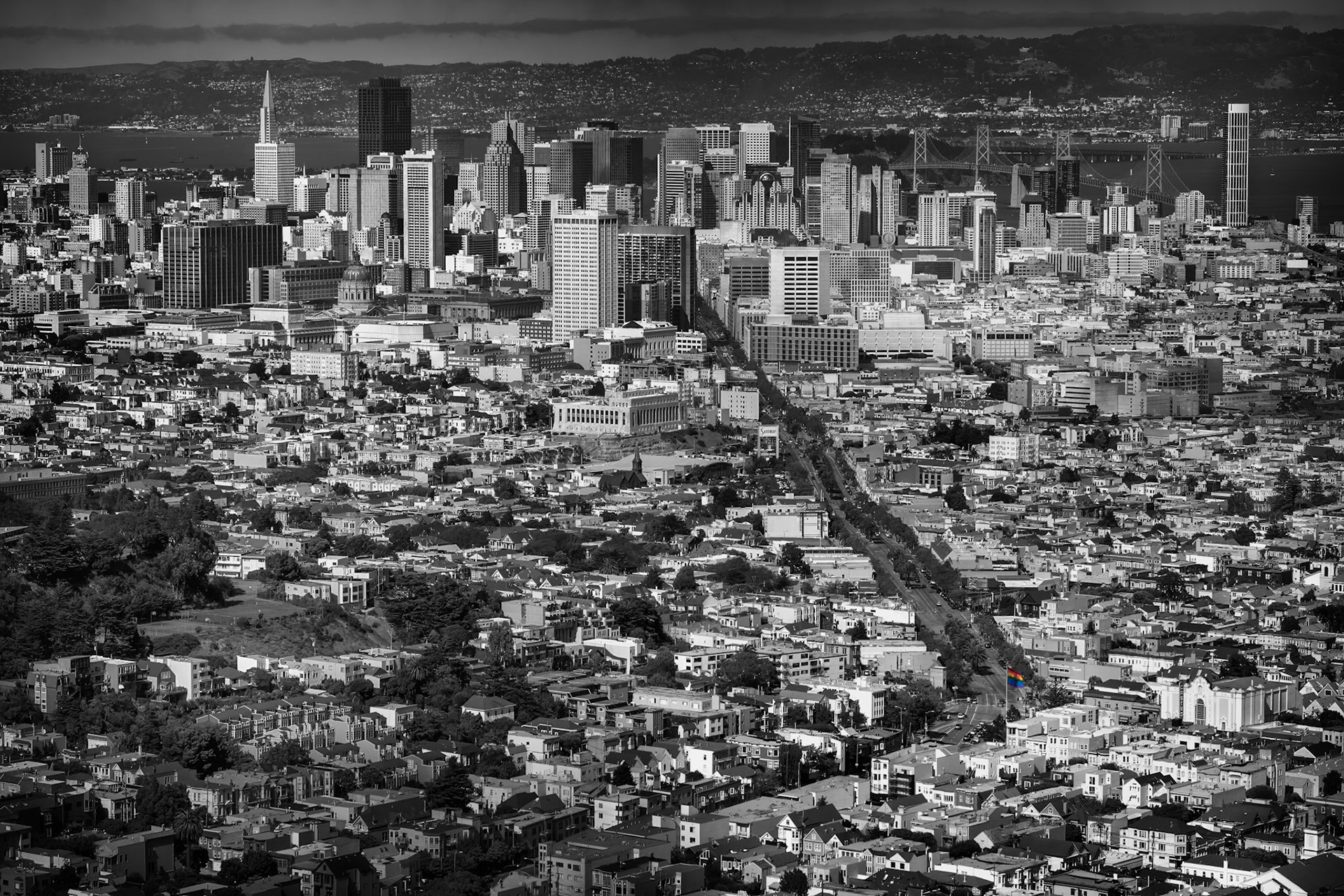 PRIDE OF CASTRO  |  A telephoto view looking down Market Street toward downtown San Francisco. The compressed perspective layers neighborhoods, commercial districts, and the city skyline into a dense urban landscape, with the Bay and distant hills forming the backdrop.Presented primarily in black and white, the image emphasizes the geometry, texture, and scale of the city. A small rainbow flag provides the only hint of color, creating a subtle focal point within the vast expanse of buildings and streets.What drew me to the scene was the way Market Street creates a visual path through the city, connecting the foreground neighborhoods to the iconic skyline beyond.