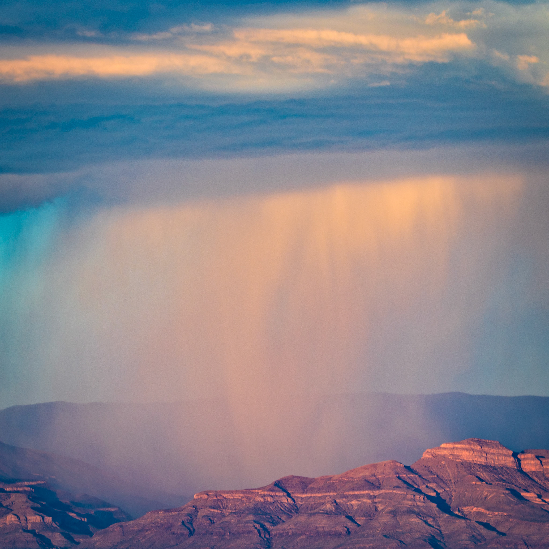 DRAIN OVER THE BASIN  |  A distant thunderstorm releases its curtains of rain across the landscape, transforming the desert sky into a study of light, atmosphere, and scale. Soft bands of illuminated precipitation descend from the clouds like brushstrokes, connecting heaven and earth in a fleeting display of nature's power.Photographed near White Sands, the scene reveals a side of the desert many never witness. While often associated with dryness and endless sun, these vast landscapes can become stages for dramatic weather, where rain falls in isolated columns and sunlight paints the storm with subtle shades of gold, lavender, and blue.The rugged ridges below provide a sense of permanence beneath a sky in constant motion. For a few brief moments, the storm becomes both spectacle and sculpture, suspended between creation and disappearance. It is a reminder that some of nature's most beautiful moments are also its most temporary, arriving quietly and vanishing just as quickly.