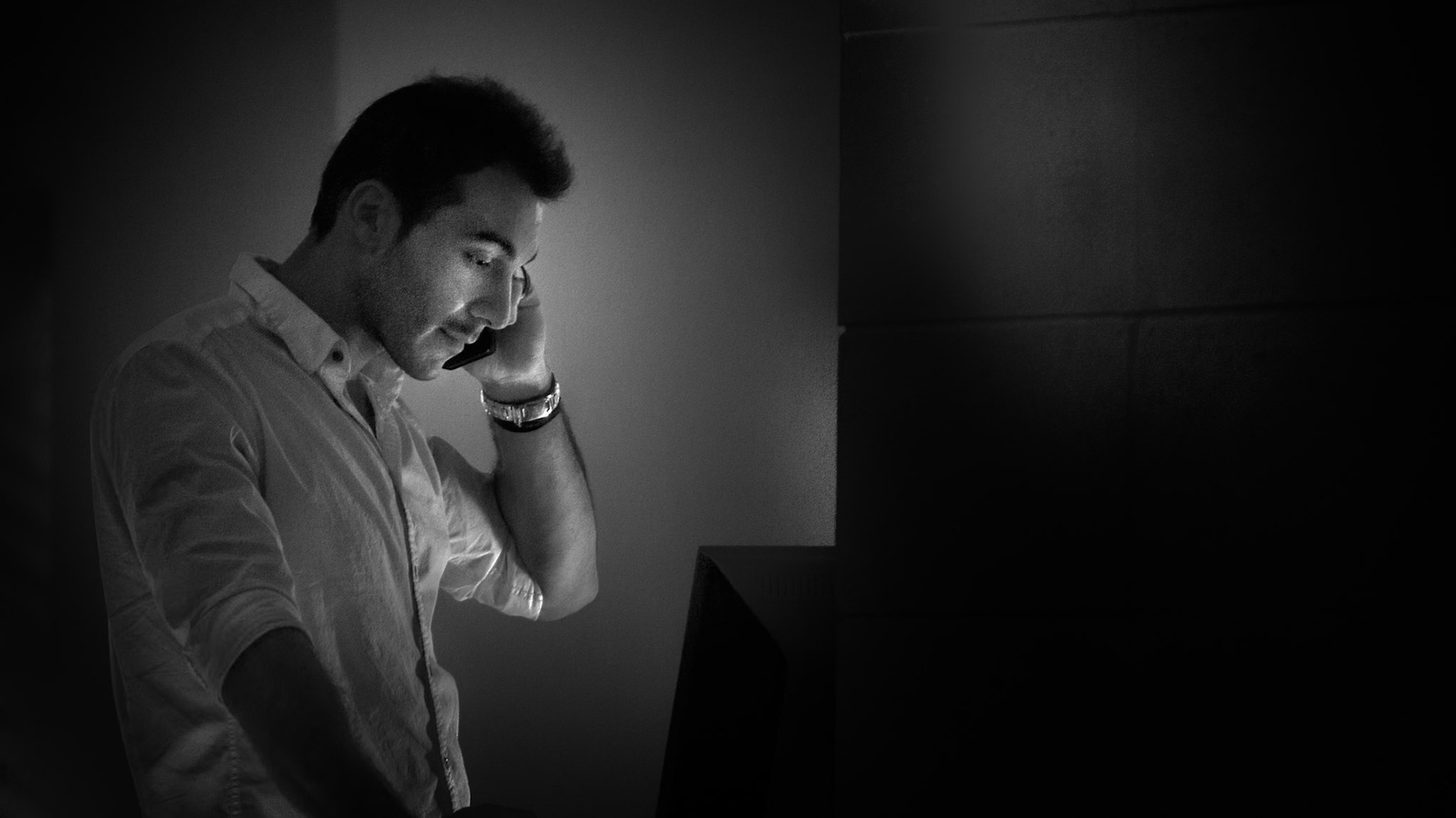 THE WEIGHT OF A CALL  |  In a world defined by constant connection, true solitude often arrives in unexpected forms. This image captures a private moment that feels both contemporary and timeless. A man stands alone in the darkness, illuminated only by the glow of a phone pressed to his ear and the faint light of a nearby screen.The black and white treatment strips away distractions, focusing attention on expression, posture, and light. Shadows dominate the frame, leaving much of the environment undefined. The viewer is left to imagine the setting, the conversation, and the story unfolding beyond the edges of the photograph.