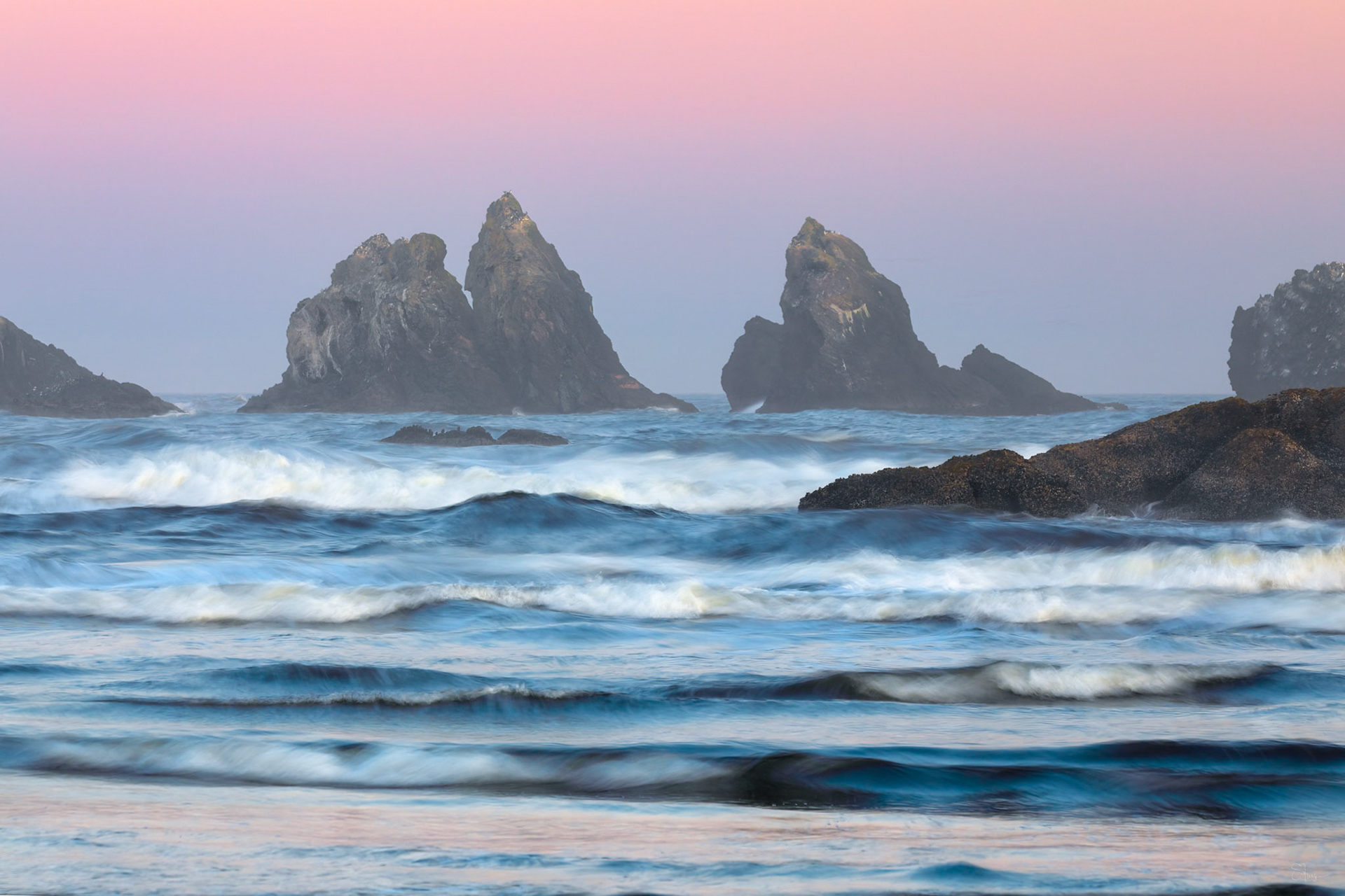 SENTINELS OF THE PACIFIC  |  As the tide rolls across Bandon Beach, waves weave delicate patterns of motion beneath sea stacks that have stood watch over the Oregon coast for centuries. The soft pastel sky and flowing water create a scene that feels both timeless and serene.Bathed in the gentle colors of twilight, the rugged rocks rise from the Pacific like ancient sentinels, their dark silhouettes contrasting beautifully with the luminous sea. The long exposure transforms the restless ocean into a graceful rhythm of texture and light.This image captures the quiet elegance of the coast, where land, sea, and sky come together in perfect harmony. It is a reminder that even in constant motion, nature can inspire a profound sense of peace.
