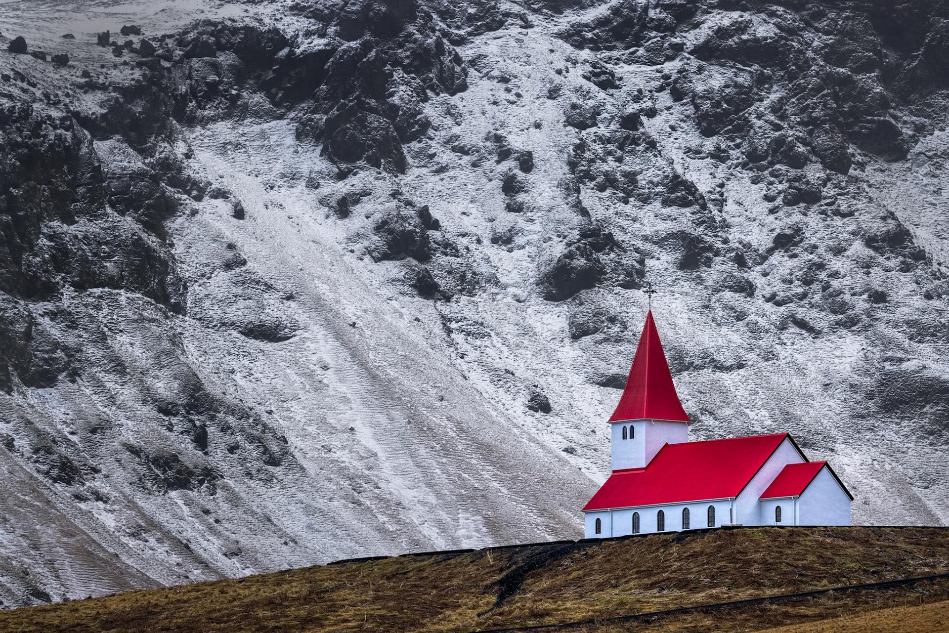 FAITH IN THE WILDERNESS  |  Nestled beneath the rugged mountains of southern Iceland, the iconic church of Vik stands as a beacon of warmth against the stark winter landscape. Its brilliant red roof draws the eye immediately, creating a striking contrast with the snow dusted cliffs that rise behind it.What makes this scene so compelling is its simplicity. There are no crowds, no distractions, just a solitary church standing resilient against the forces of nature. The surrounding landscape feels wild and untamed, while the church offers a sense of shelter, hope, and human presence.Traveling through Iceland in winter often feels like stepping into another world. This image captures one of those moments when color, light, and place come together to tell a story of endurance, faith, and quiet beauty at the edge of the North Atlantic.