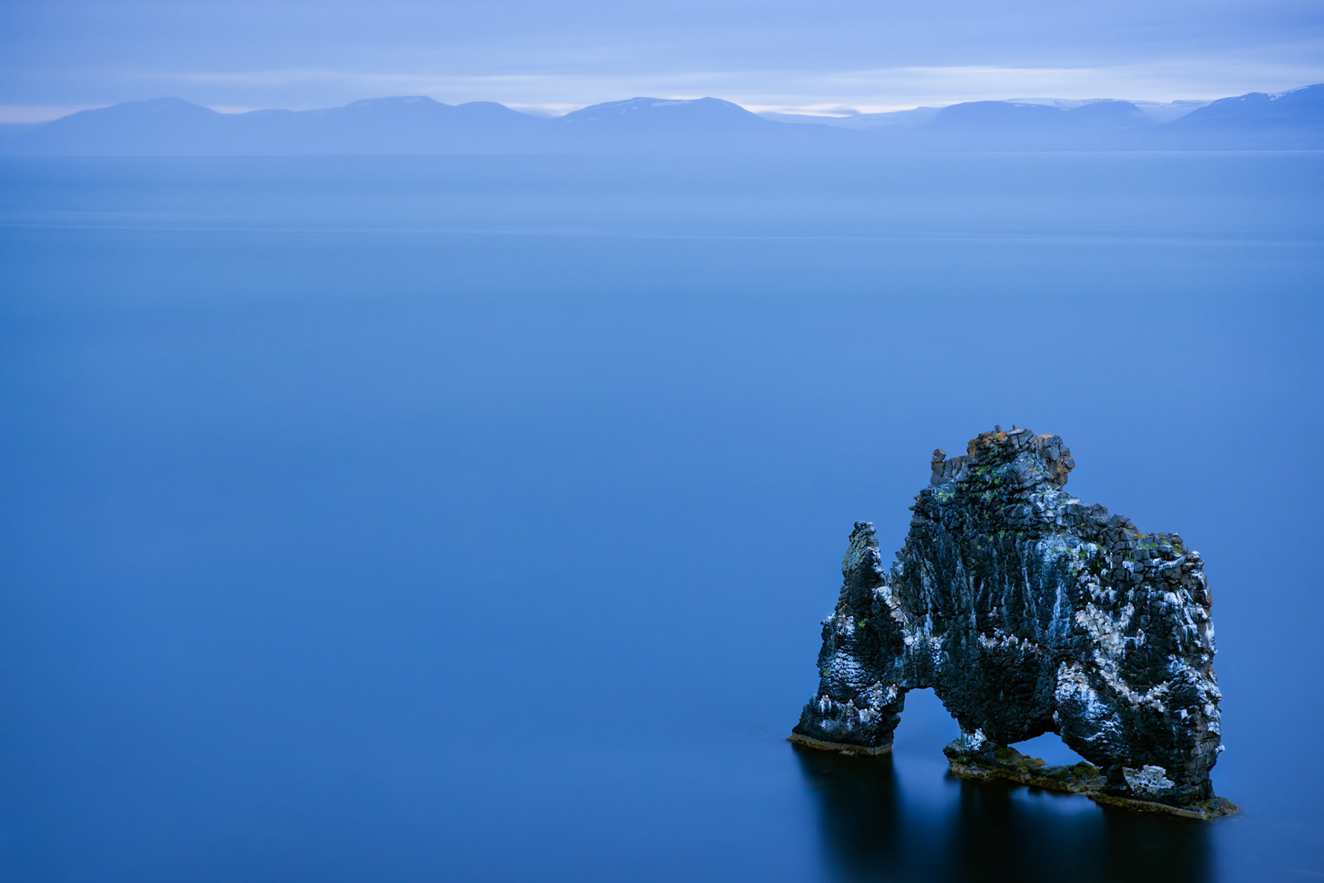 THE STONE GIANT  |  Rising from the still waters of northwest Iceland, Hvítserkur stands alone against the vastness of sea and sky. Shaped by centuries of wind and waves, the basalt formation has inspired legends, with many seeing the silhouette of a dragon, a rhinoceros, or a mythical creature frozen in stone.
