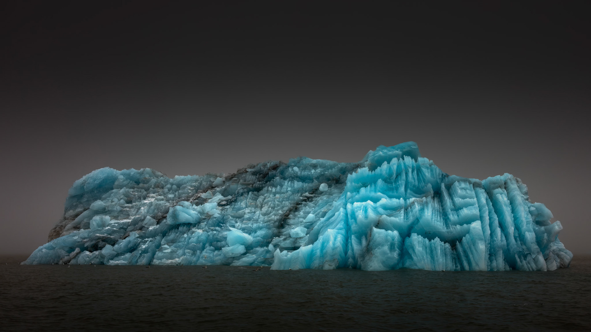 FROZEN TITAN  |  Emerging from the Arctic fog, this iceberg reveals a world hidden beneath the surface. Layers of compressed snow and ancient ice have been sculpted by wind, waves, and time into a cathedral of crystalline blue, its textures recording centuries of frozen history.