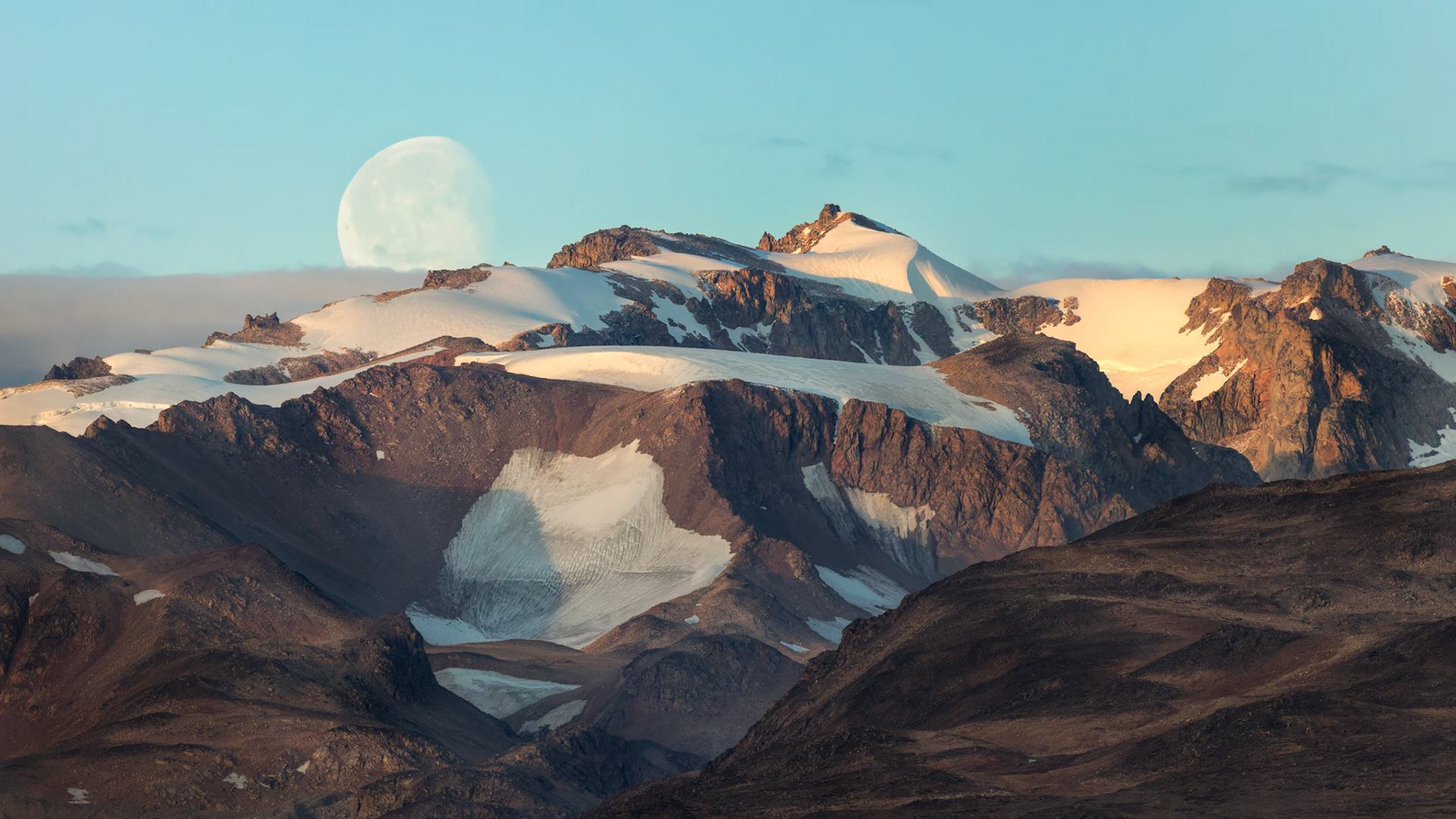 MOONRISE OVER SCORESBY SOUND  |  A pale moon rises behind the glaciated mountains of Scoresby Sound, Greenland, as the last warm light of day touches the rugged peaks. The contrast of soft lunar light, snow, and dark volcanic rock gives the landscape an almost otherworldly stillness.