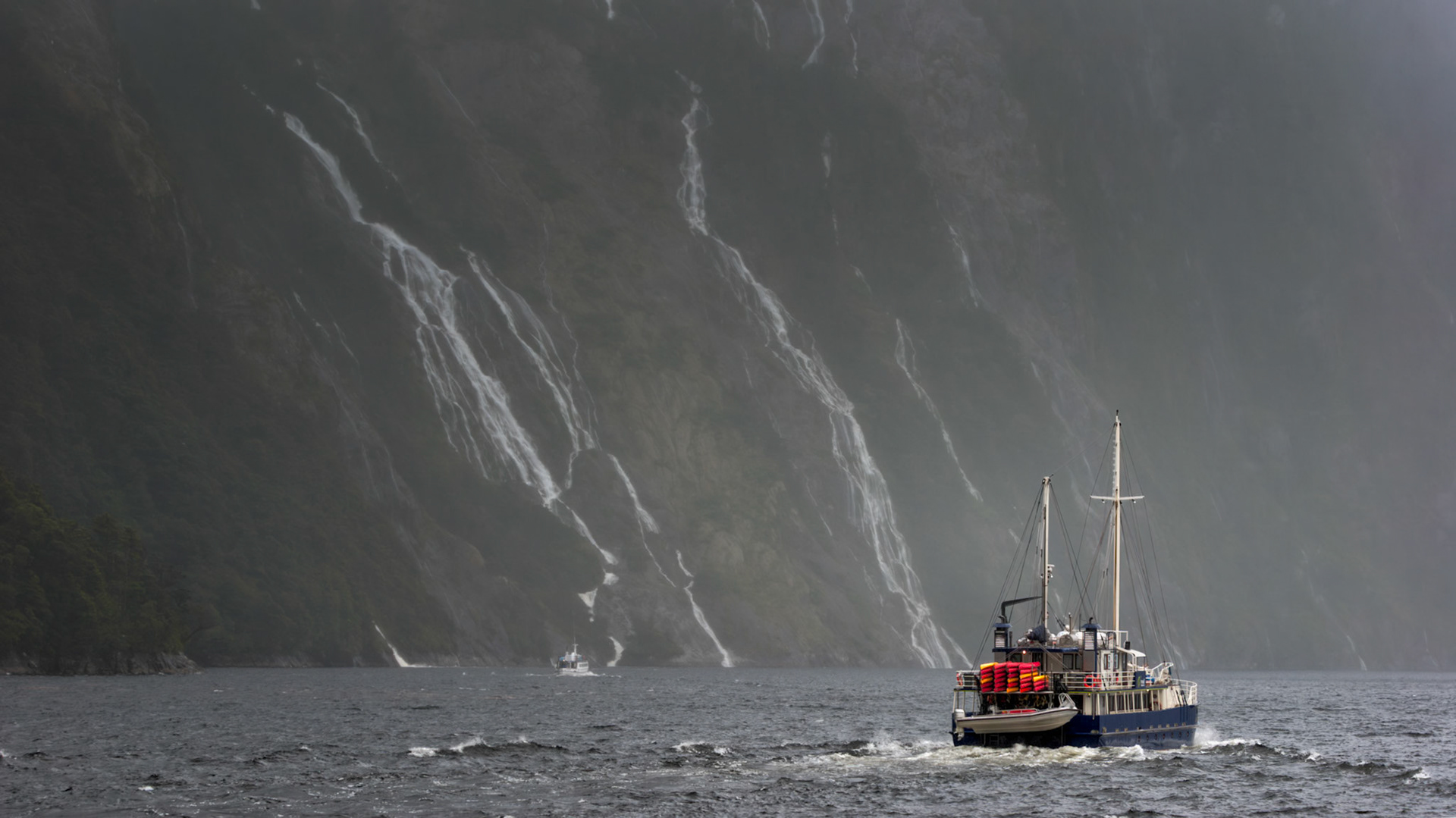 INTO THE MIST  |  Few places make you feel as small as Milford Sound. As our vessel moved across the dark water, it seemed substantial enough until it passed beneath cliffs that rise thousands of feet almost straight from the sea. Suddenly, the boat became little more than a speck in a landscape shaped long before humans ever arrived.Waterfalls spilled from the sheer rock walls like silver threads, disappearing into the mist that constantly drifts through the fiord. Rain, glaciers, and time have carved this remarkable place over millions of years, creating a landscape that feels less like New Zealand and more like a glimpse into a prehistoric world.Standing here, it was impossible not to feel humbled by the scale of nature. The boat provides perspective, a reminder that in places like Milford Sound, we are merely visitors passing briefly through a landscape that has endured for ages and will continue long after we are gone.