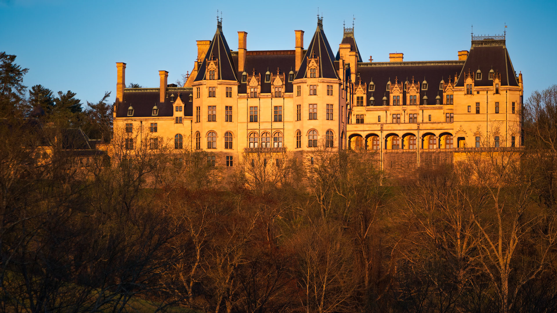 EVENING AT THE ESTATE  |  As the sun nears the horizon, warm golden light washes across the limestone façade of Biltmore House, accentuating the architectural details and steeply pitched roofs that define America's largest privately owned home.Completed in 1895 for George Vanderbilt, the estate was inspired by the grand châteaux of France's Loire Valley. From this vantage point, the mansion rises above the surrounding landscape, its formal design contrasting with the natural woodland below.The low-angle sunlight briefly transforms the building, illuminating the stonework while long shadows begin to settle across the grounds. The result is a portrait of Biltmore at one of the most photogenic moments of the day, when architecture and light come together to reveal the estate's enduring elegance.