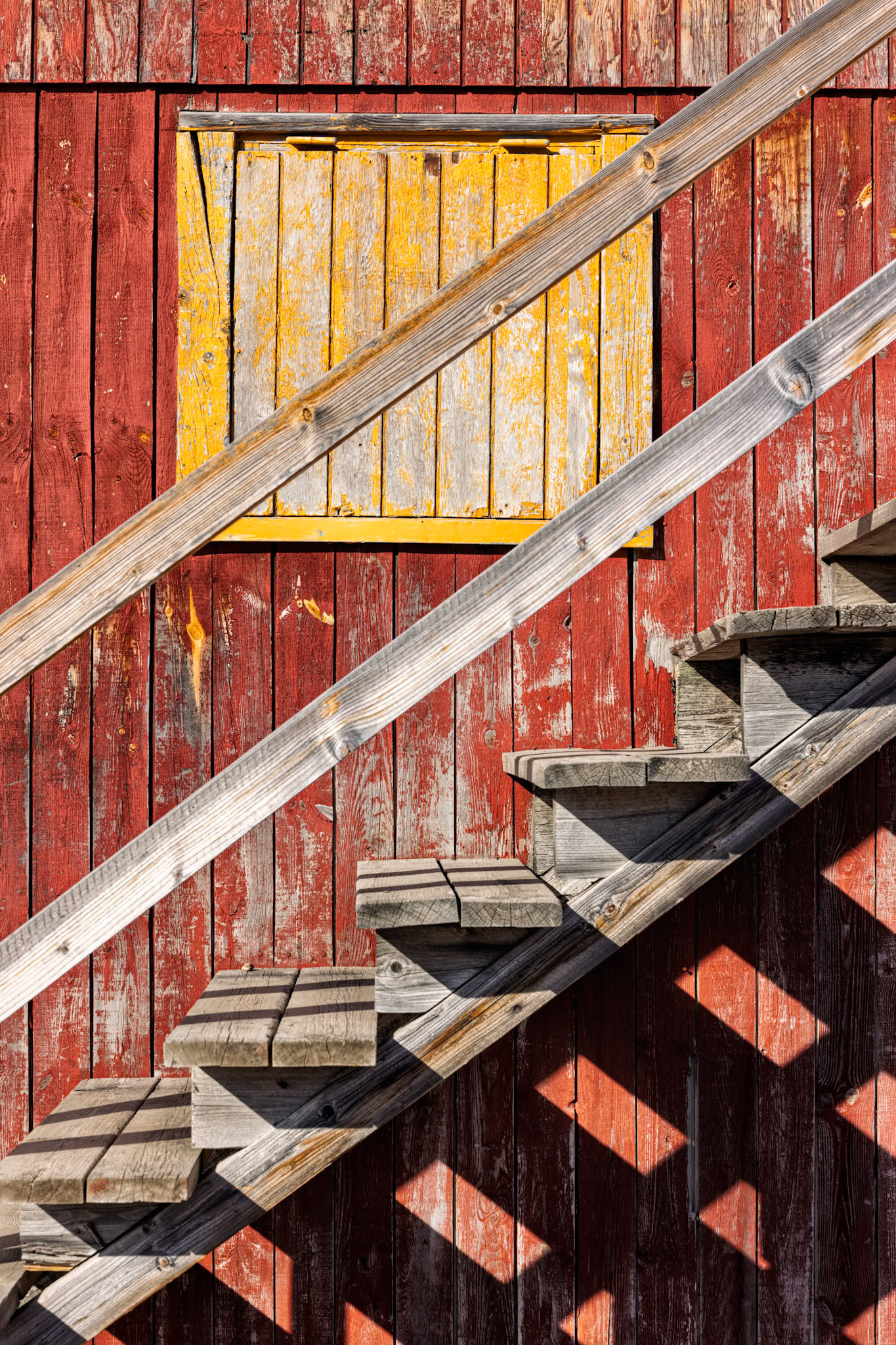 ARCTIC GEOMETRY  |  Weathered red siding, a sunlit yellow shutter, and the strong diagonal lines of a wooden staircase turn a simple Greenland building into a study of color, texture, and shape. Hard Arctic light creates a second layer of geometry through the repeating shadows, giving the composition an almost abstract quality.