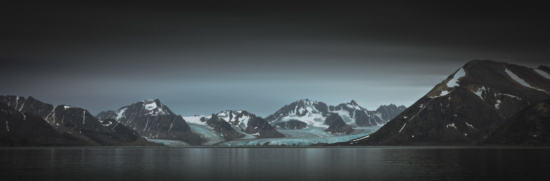 WHERE ICE MEETS SILENCE | Ancient glaciers spill between rugged mountains into the still waters of Svalbard, where light, ice, and stone exist in quiet balance. The scene invites reflection on the enduring beauty of landscapes shaped over thousands of years.