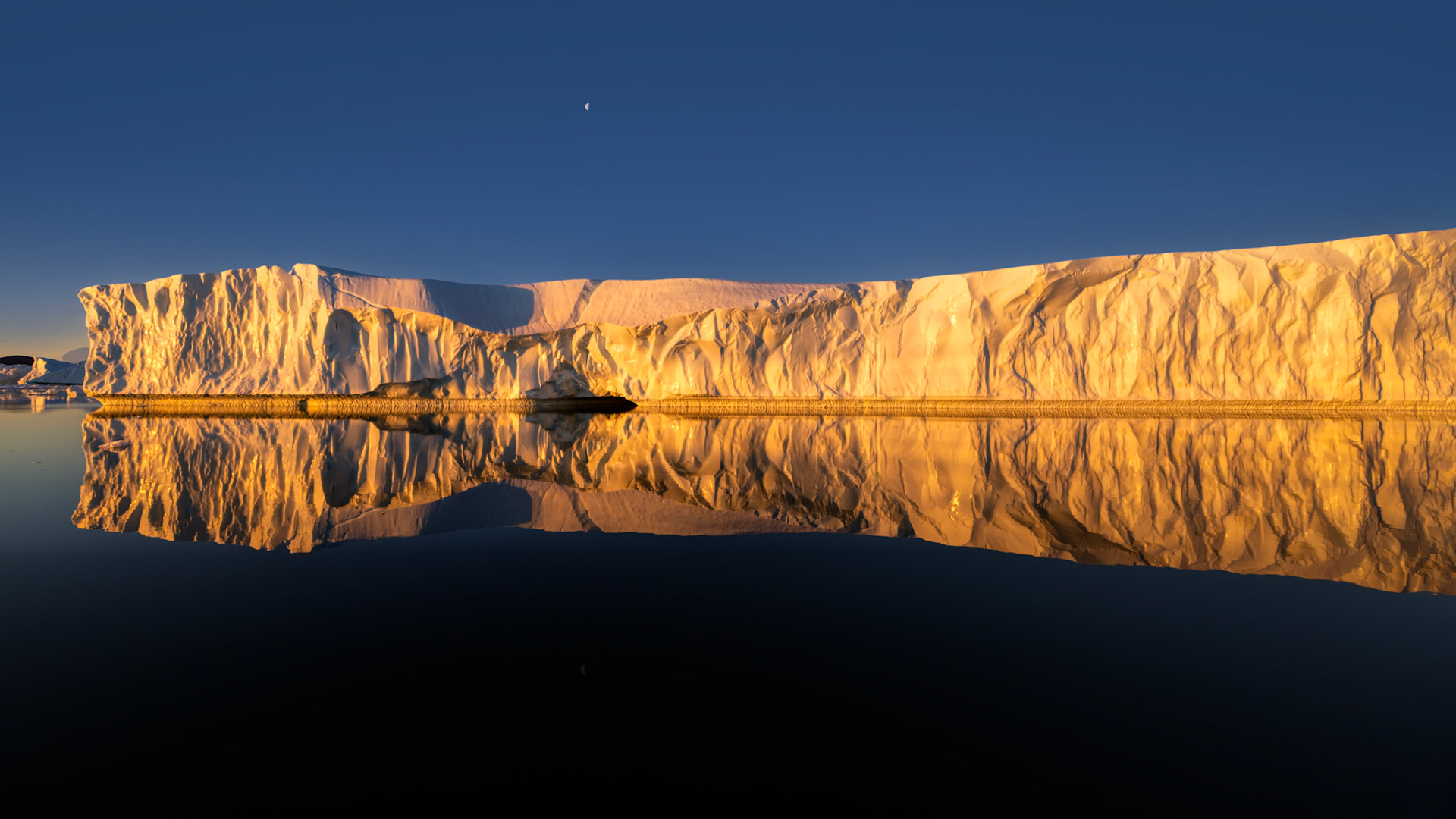 WHEN ICE BURNS GOLD  |  In Greenland's summer twilight, the sun briefly slips beneath the clouds and transforms an ancient iceberg into a wall of gold. For a few fleeting moments, ice that has drifted through Arctic waters for centuries glows with warmth, creating a striking contrast between the frozen landscape and the light that brings it to life.The stillness was extraordinary. The water became a perfect mirror, doubling the iceberg's intricate textures and allowing the reflection to stretch endlessly into the darkness below. Even the small moon hanging overhead seemed to pause and watch the spectacle unfold.This is one of those rare moments photographers dream about. The light lasted only minutes, but it revealed a side of the Arctic that few people imagine. Not cold and distant, but luminous, peaceful, and unexpectedly radiant.